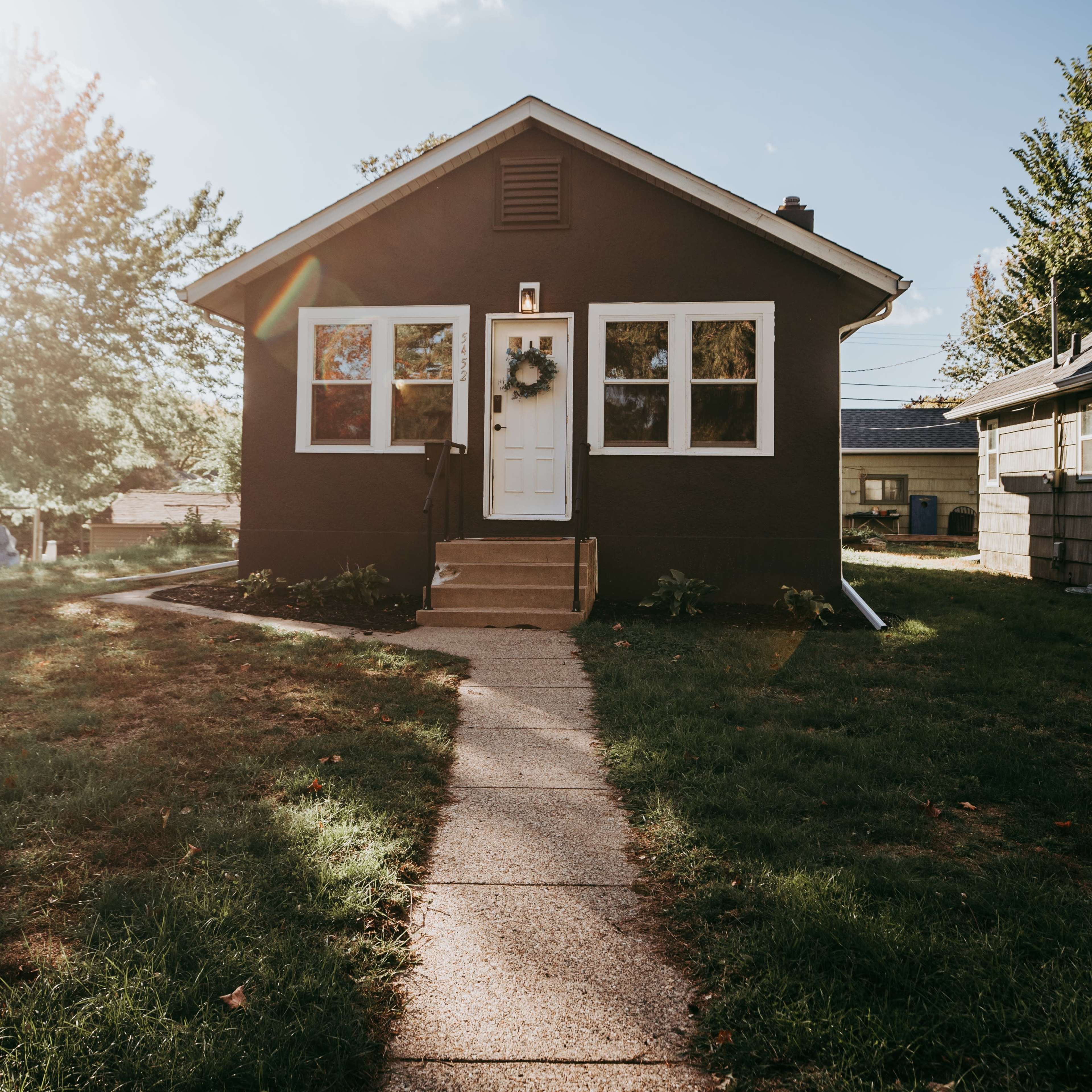 A small dark-colored house with a white front door and a wreath is surrounded by a lawn and a concrete pathway.