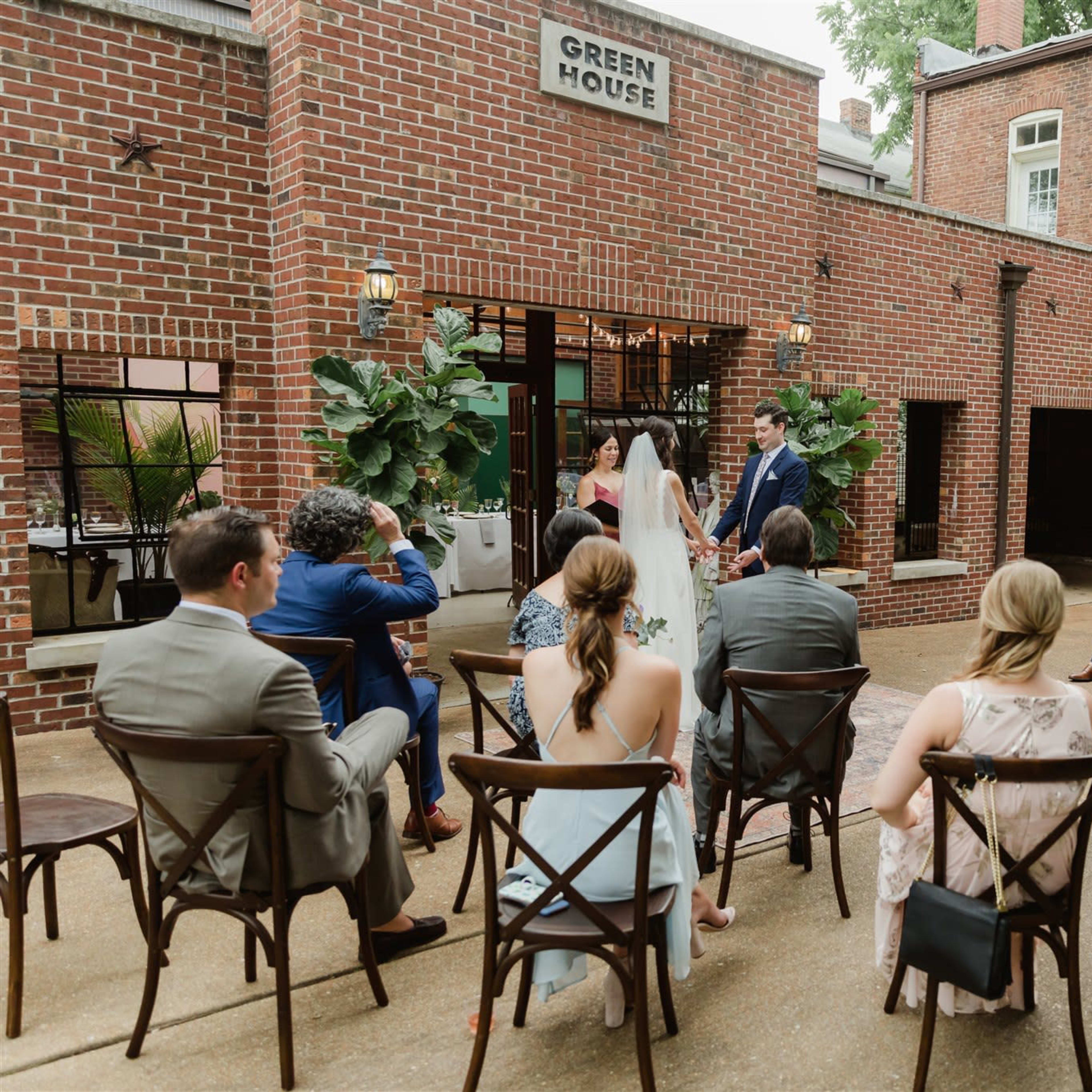 A couple stands at the front of a courtyard, exchanging vows while guests seated in rows of chairs watch.