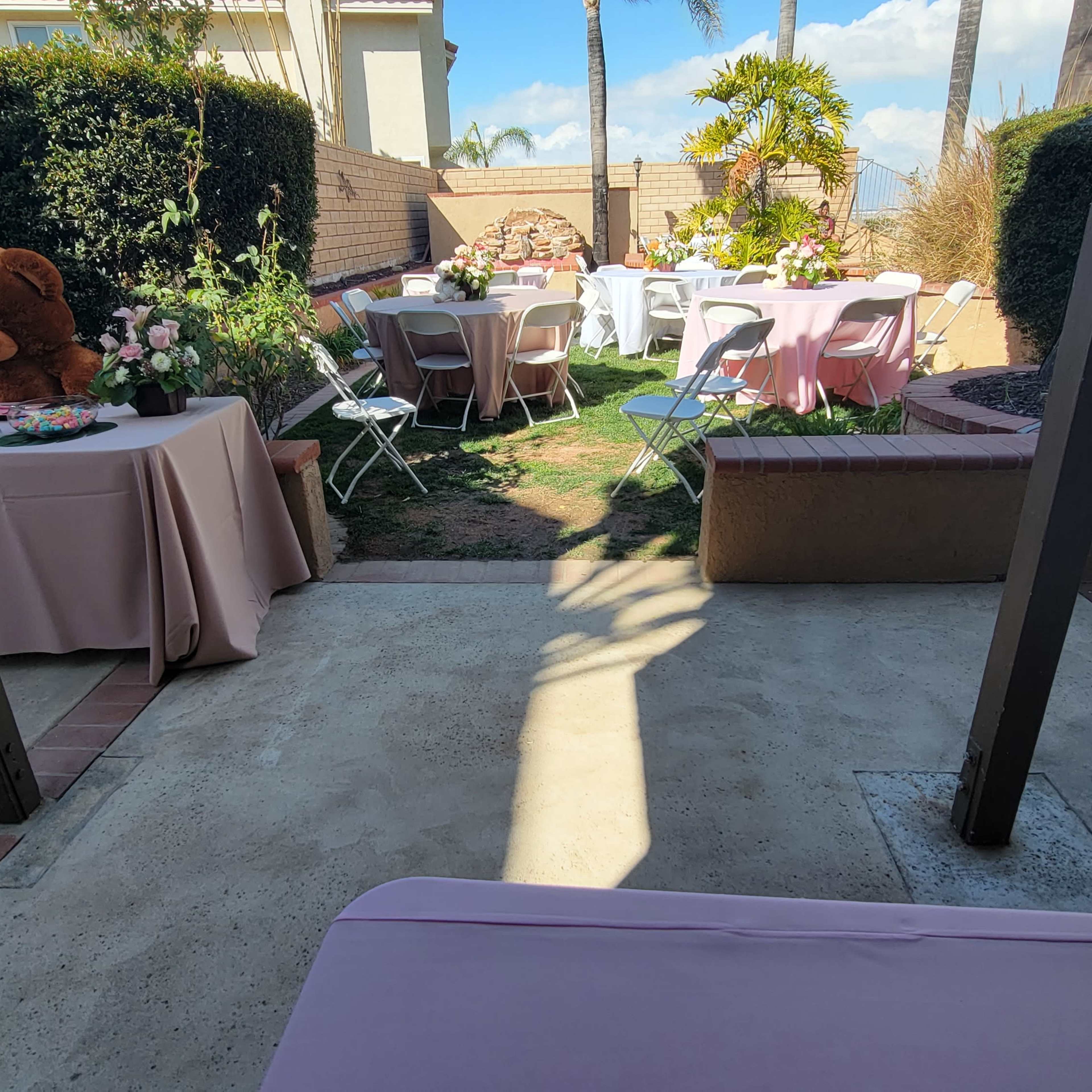 The scene shows an outdoor patio set up for a gathering, featuring tables covered with pink tablecloths, a large teddy bear, floral centerpieces, and a lawn area with palm trees in the background.