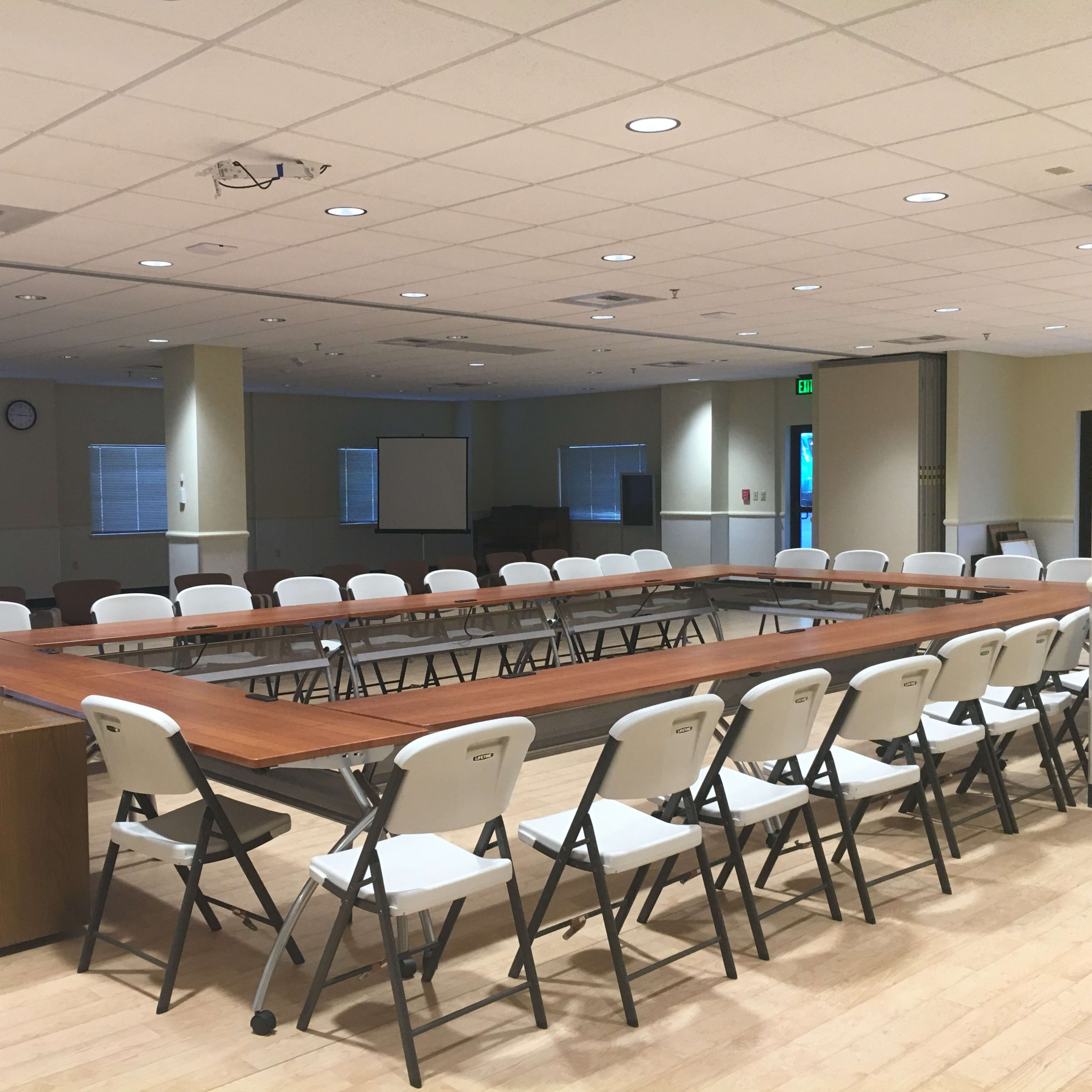 A meeting room with a large rectangular table surrounded by folding chairs and large windows.