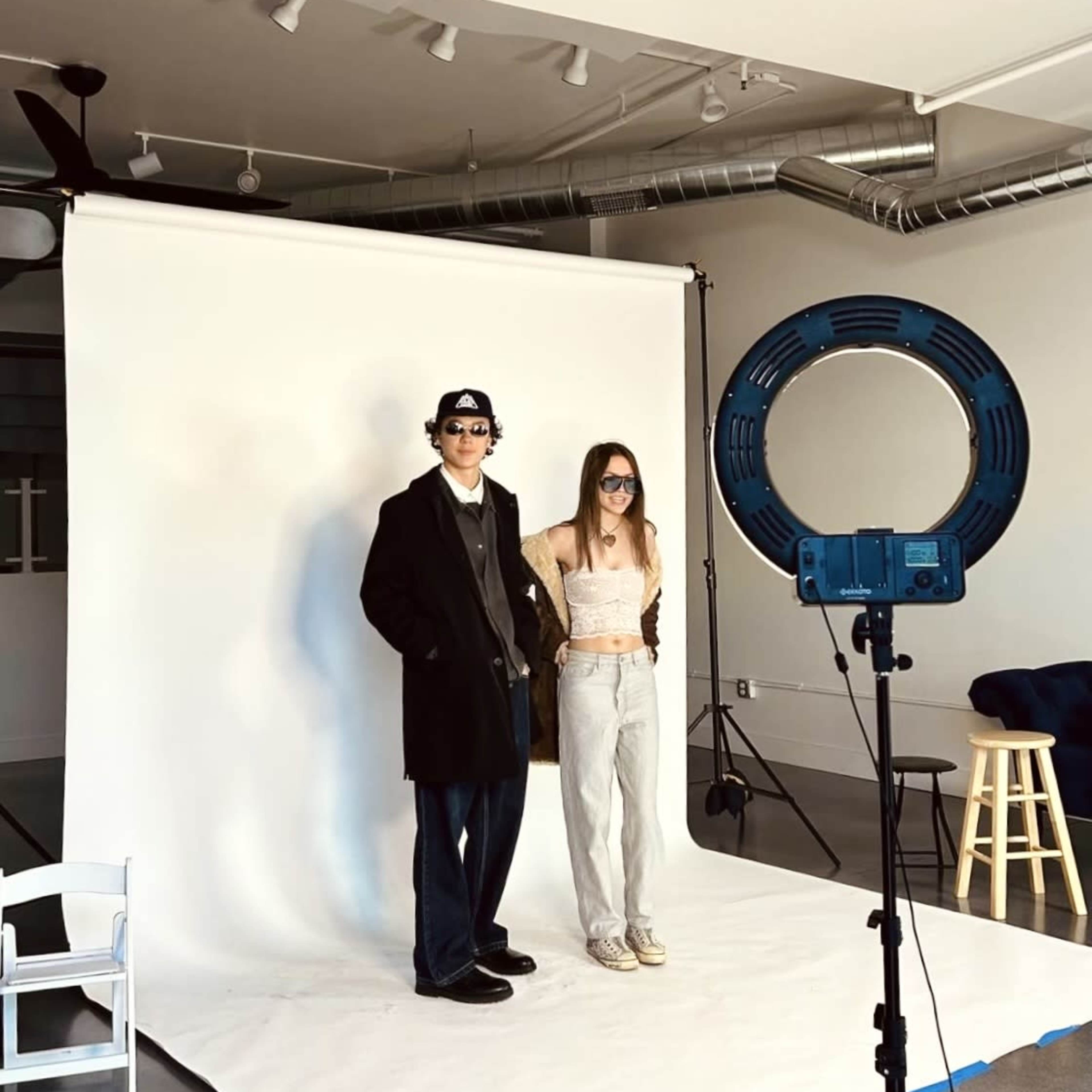 A man and a woman pose for a photo in a studio setting with a white backdrop and a ring light.