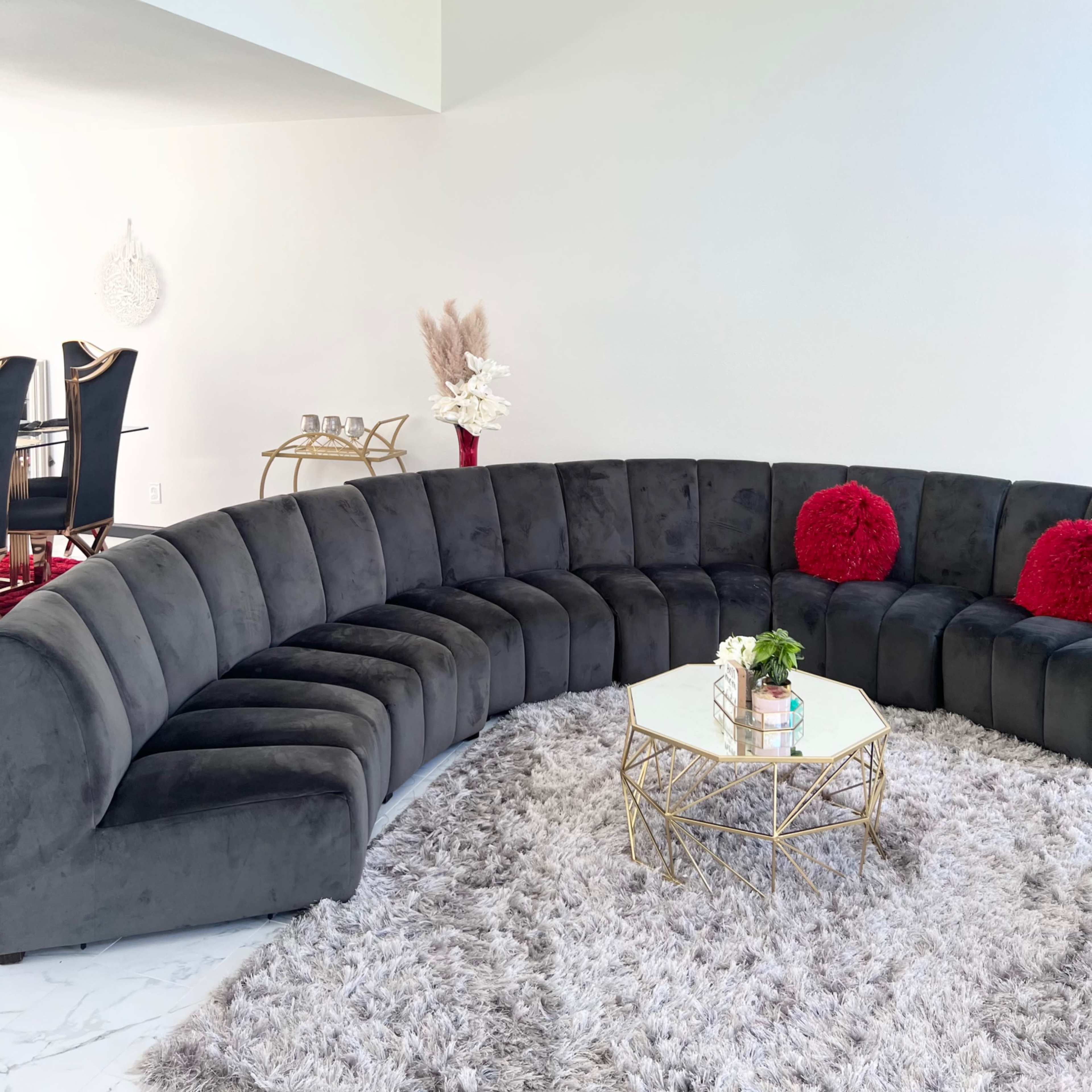 A curved black sofa with red pillows is arranged around a gold coffee table on a fluffy gray rug in a well-lit room.