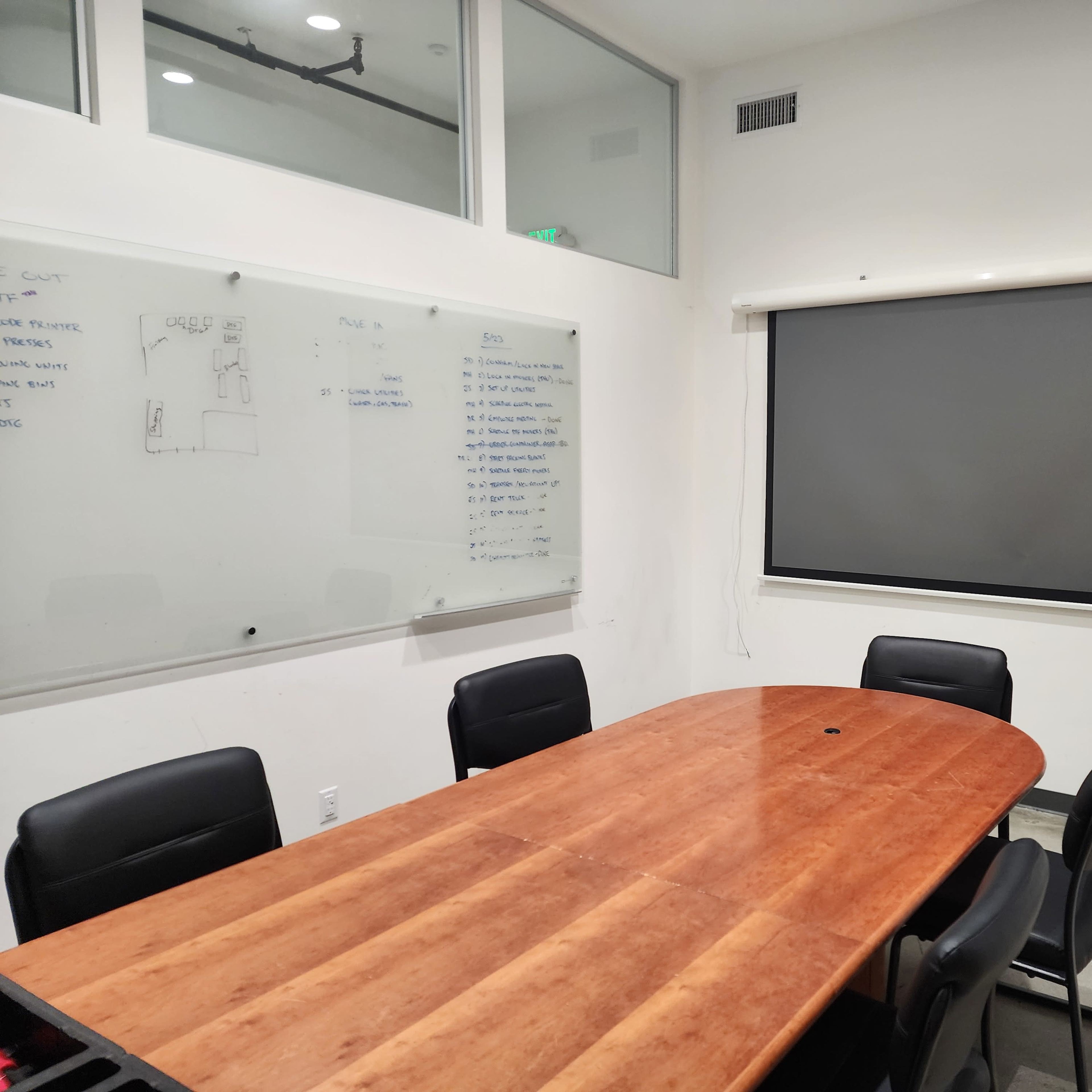 A conference room features a large wooden table surrounded by black chairs, a whiteboard on one wall, and a projection screen on another wall.