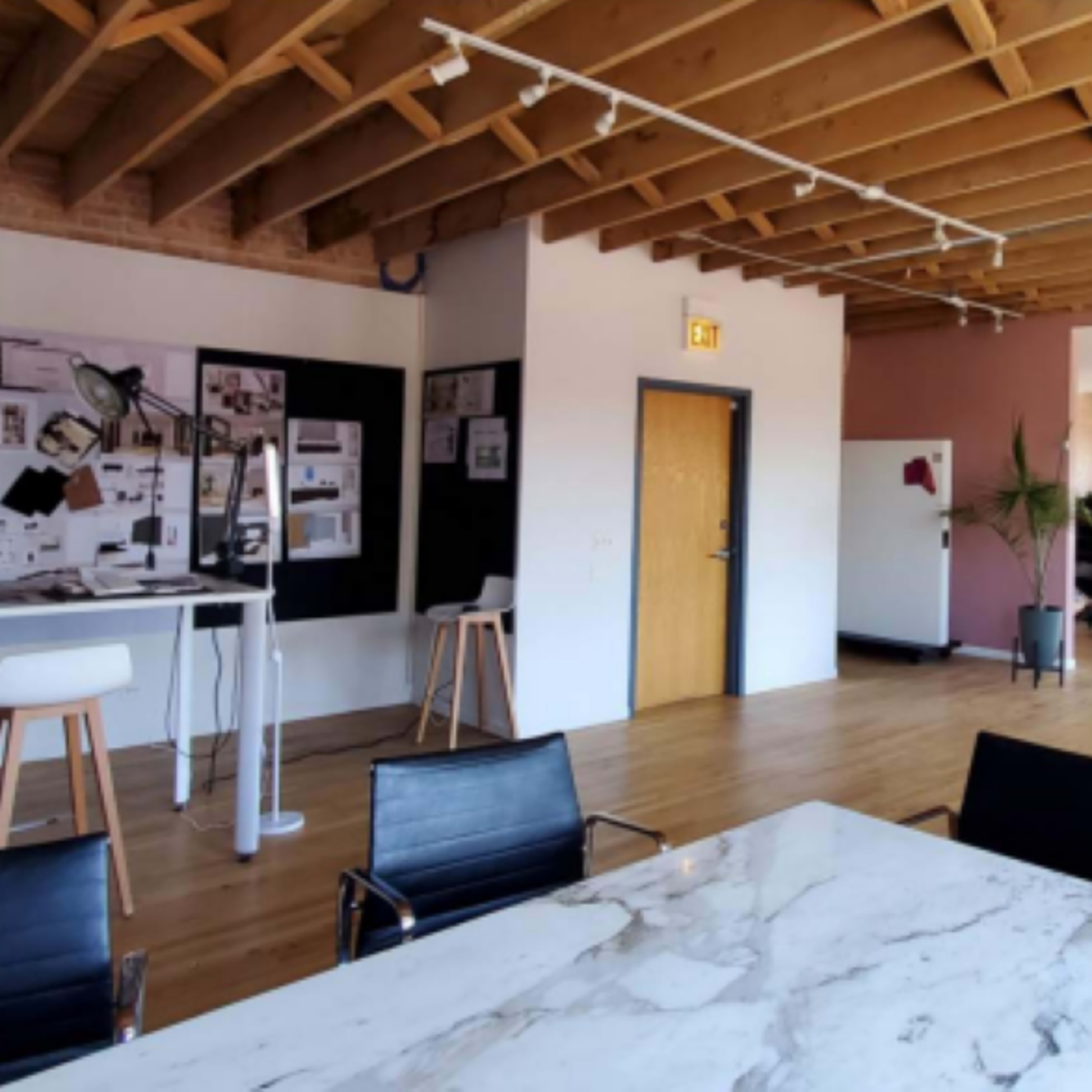 A spacious office interior with wooden flooring, exposed beams, and plants, featuring a large table, chairs, and workstations in the background.