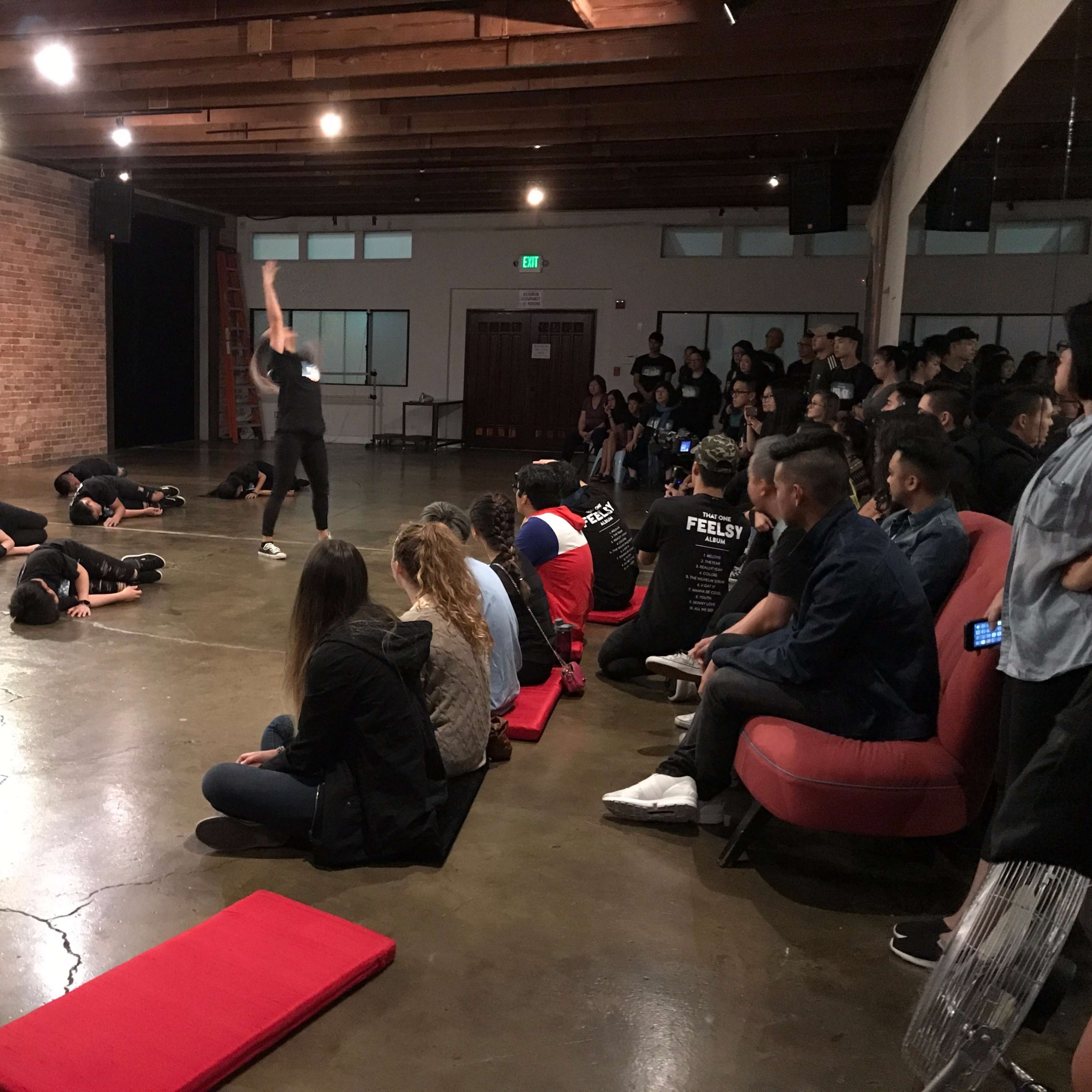 A performance scene takes place in a dimly lit space with a group of dancers rehearsing on the floor, while an audience watches from nearby seating.