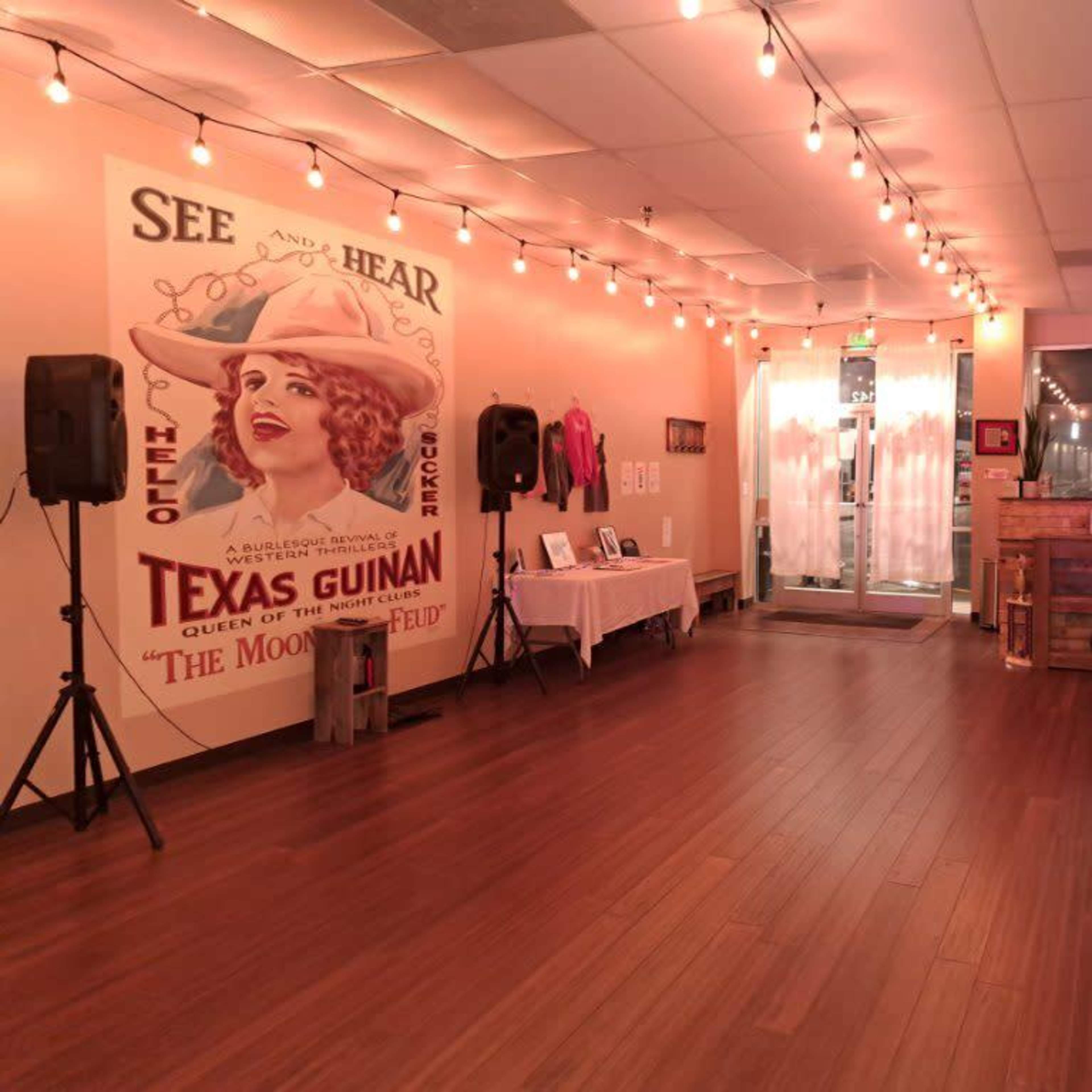 An interior space featuring wooden flooring, a large vintage poster of Texas Guinan on the wall, and a wooden reception desk near a glass entrance with string lights overhead.