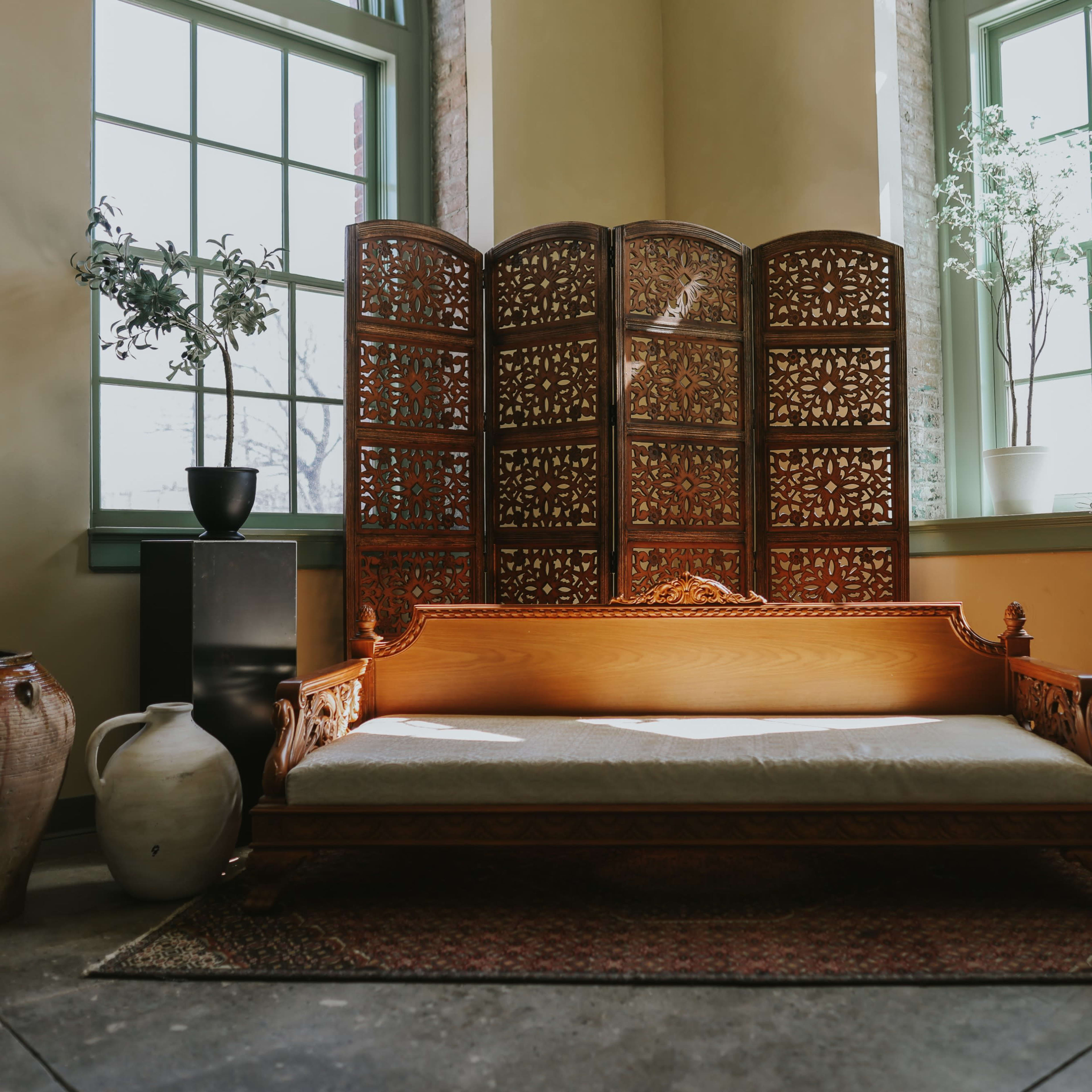 A wooden sofa with intricate carvings set against a backdrop of large windows and a decorative room divider, surrounded by potted plants and various decorative items.