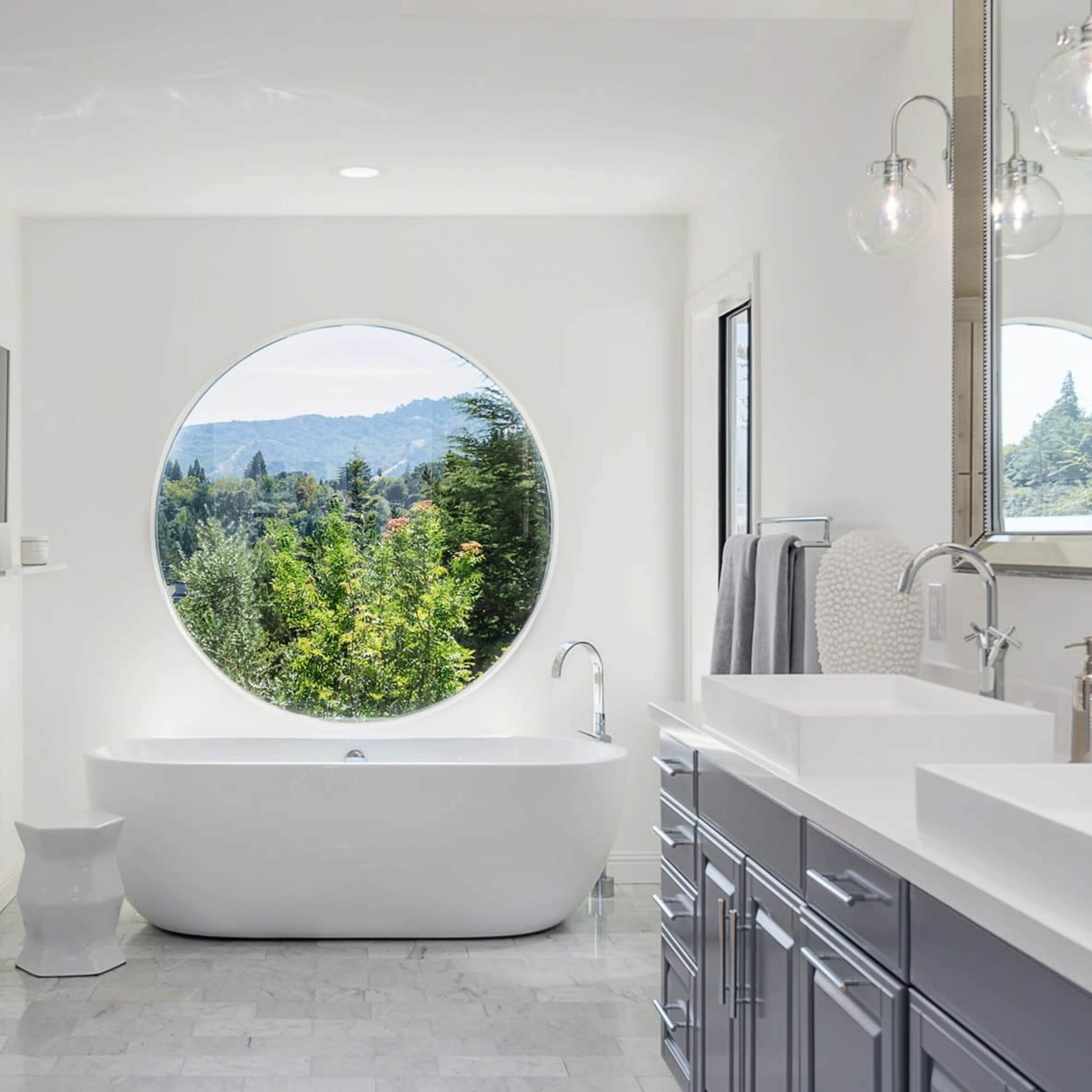 The bathroom features a freestanding white tub next to a large circular window that offers a view of outdoor greenery.