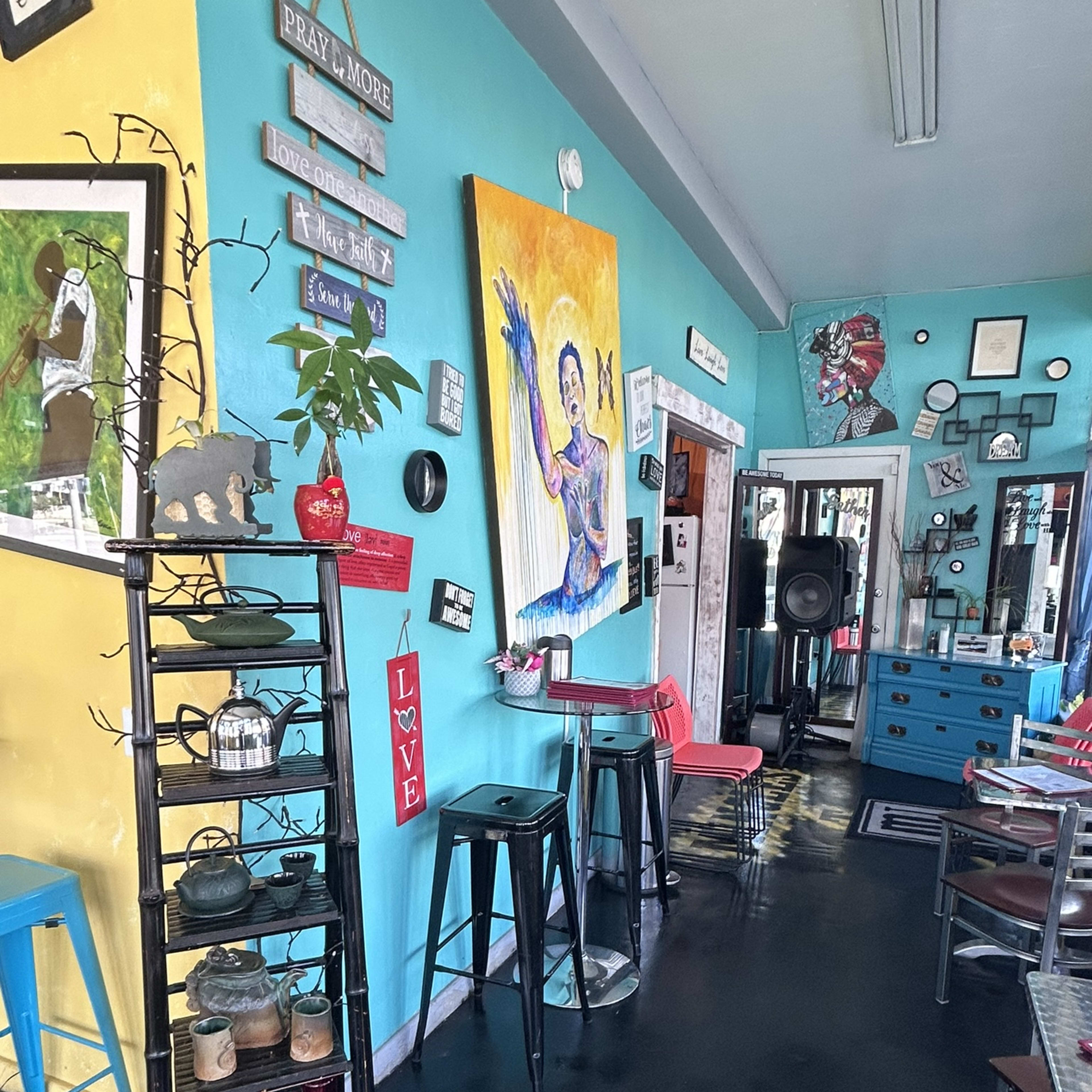 A colorful café interior with turquoise and yellow walls, featuring a vibrant mural, wooden shelves with décor, and various seating arrangements.