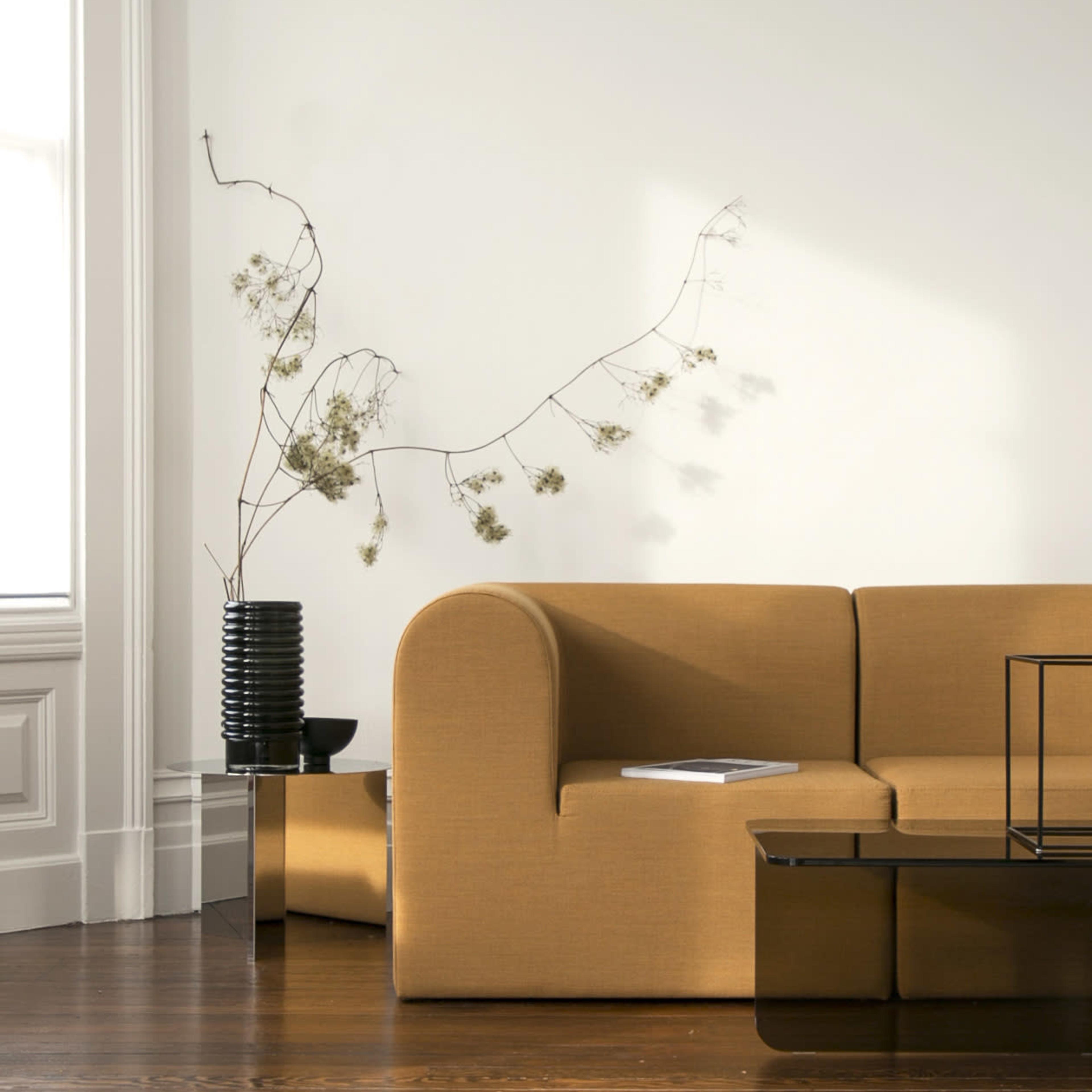A modern living room features a brown sofa, a black decorative vase with dried flowers, and a glass coffee table against a light-colored wall.
