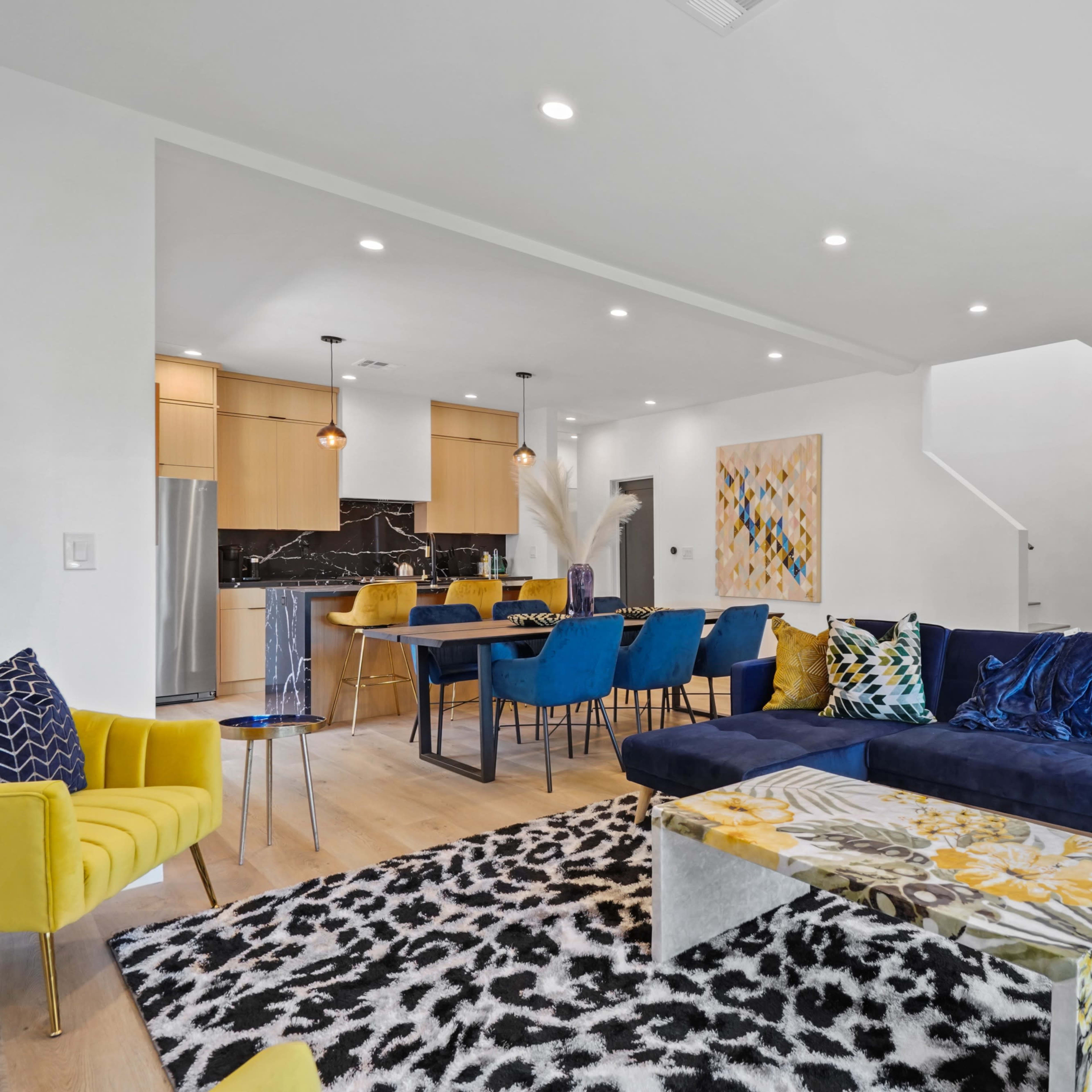 A modern living and dining area featuring a navy blue couch, colorful chairs, a patterned rug, and a contemporary kitchen in the background.