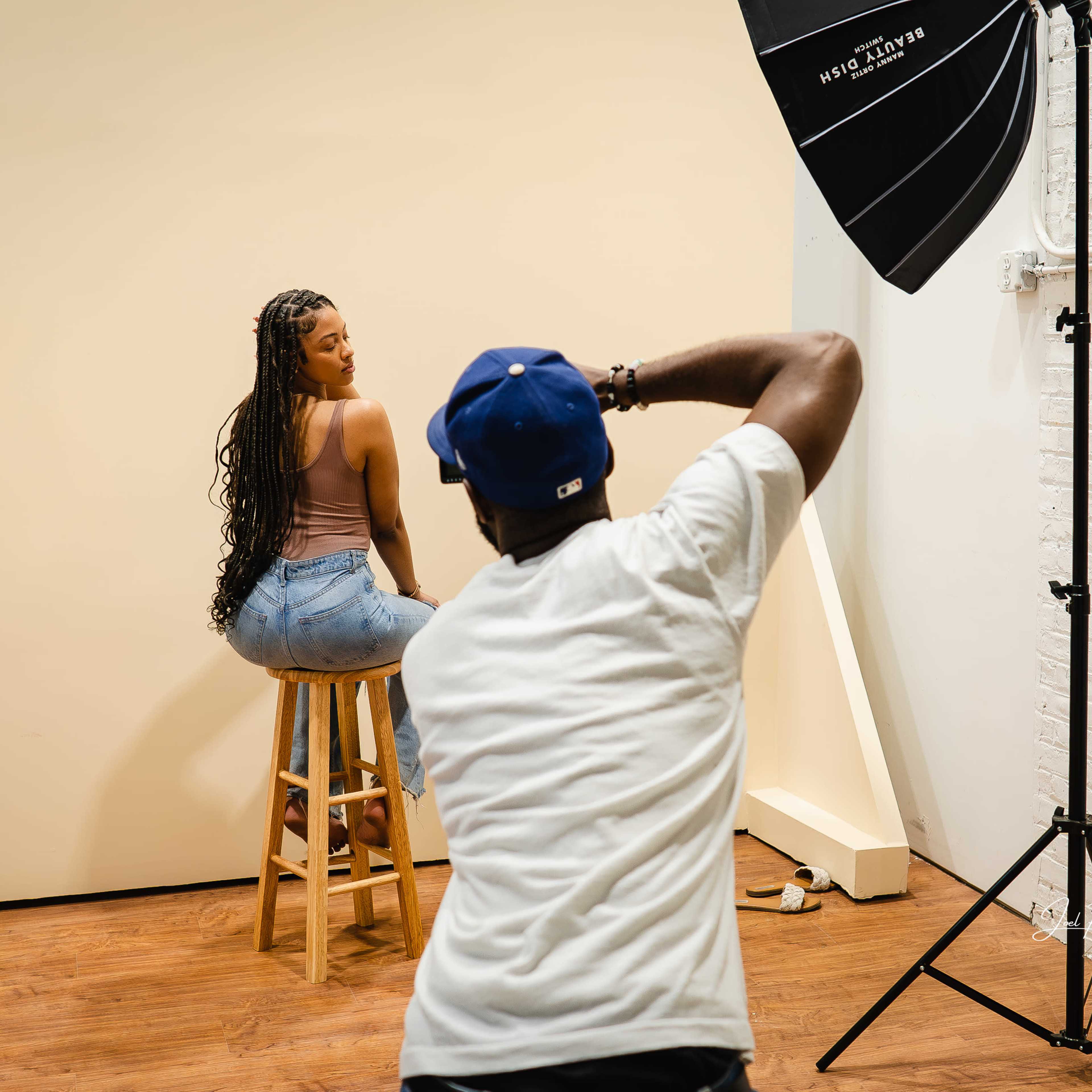 A photographer captures a model posing on a stool in a studio with a neutral background and lighting setup.