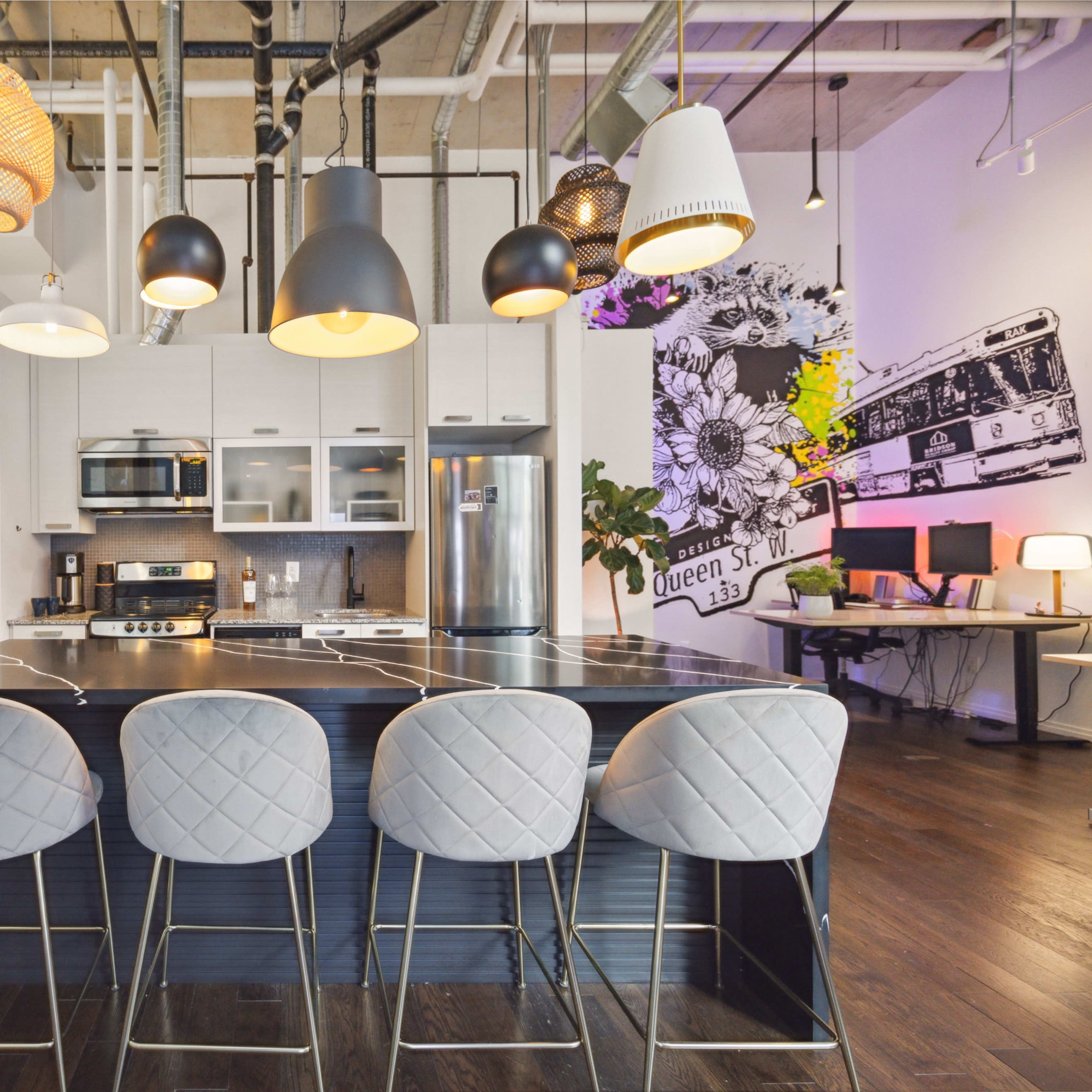 A modern kitchen features a central island with four barstools, pendant lighting, and a mix of appliances against a backdrop of vibrant wall art.