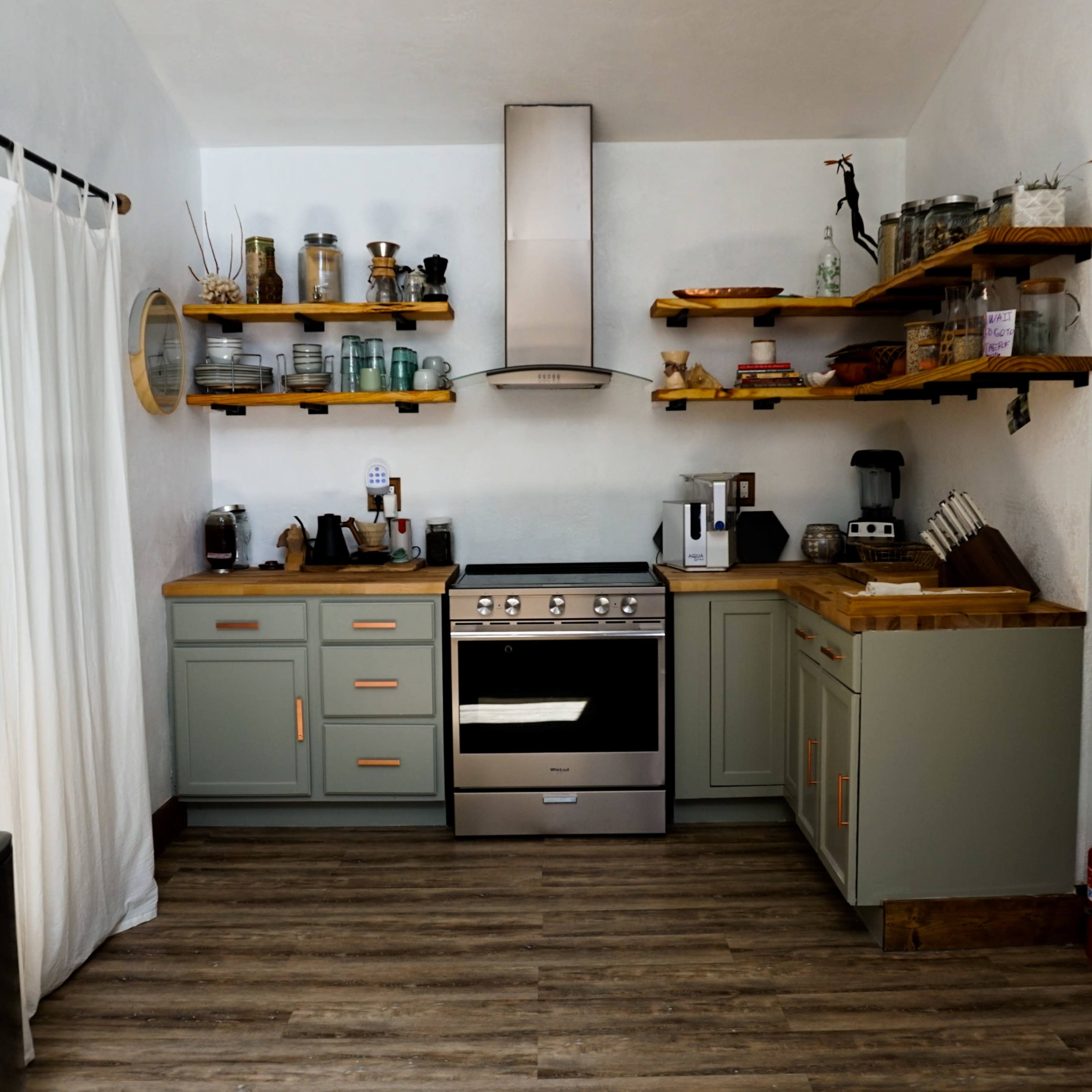 The kitchen features gray cabinets, wooden countertops, and shelves holding various kitchen items, with stainless steel appliances and a large window.