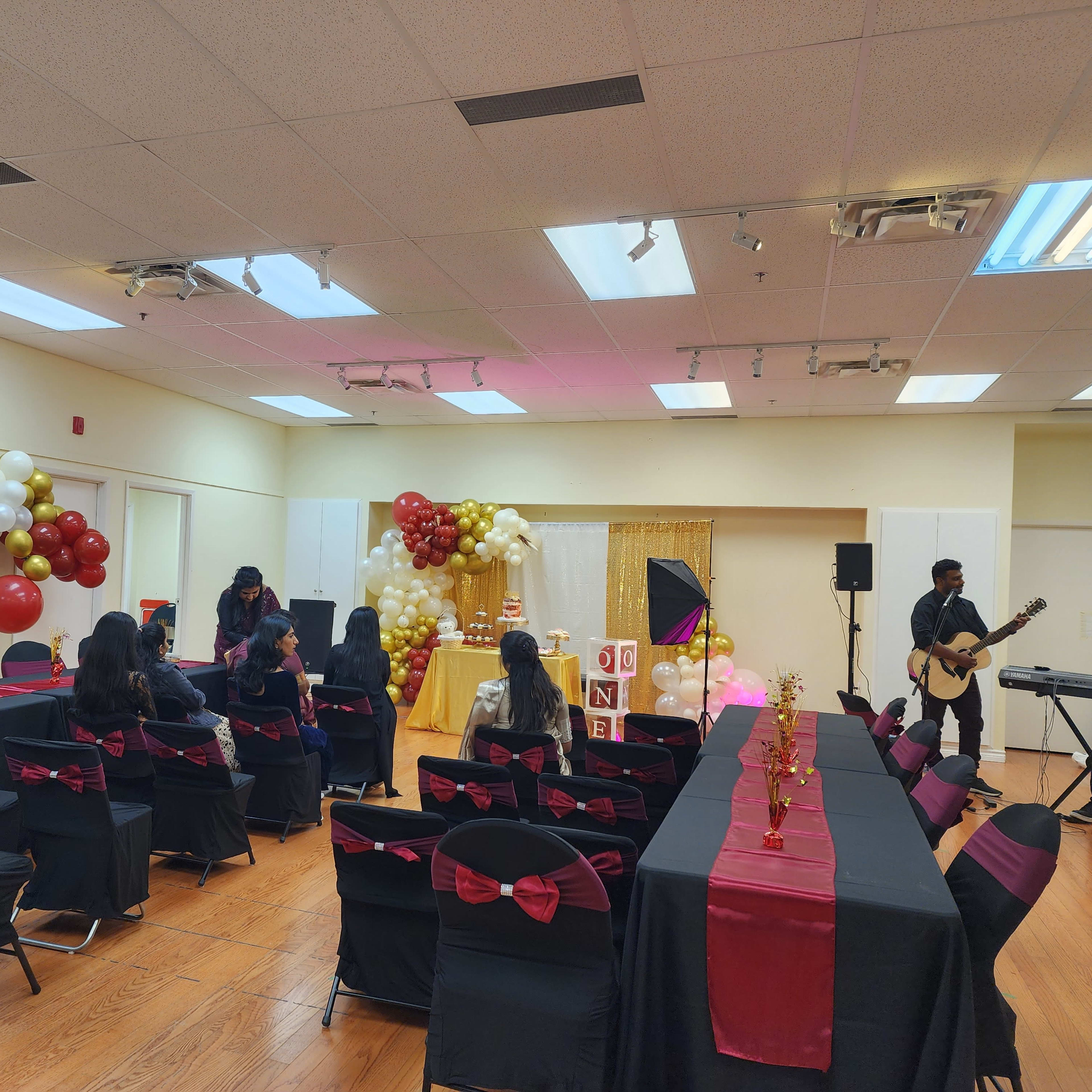 A decorated room with black tables and chairs featuring red bows, gold and white balloons, and a small stage set up for performers.