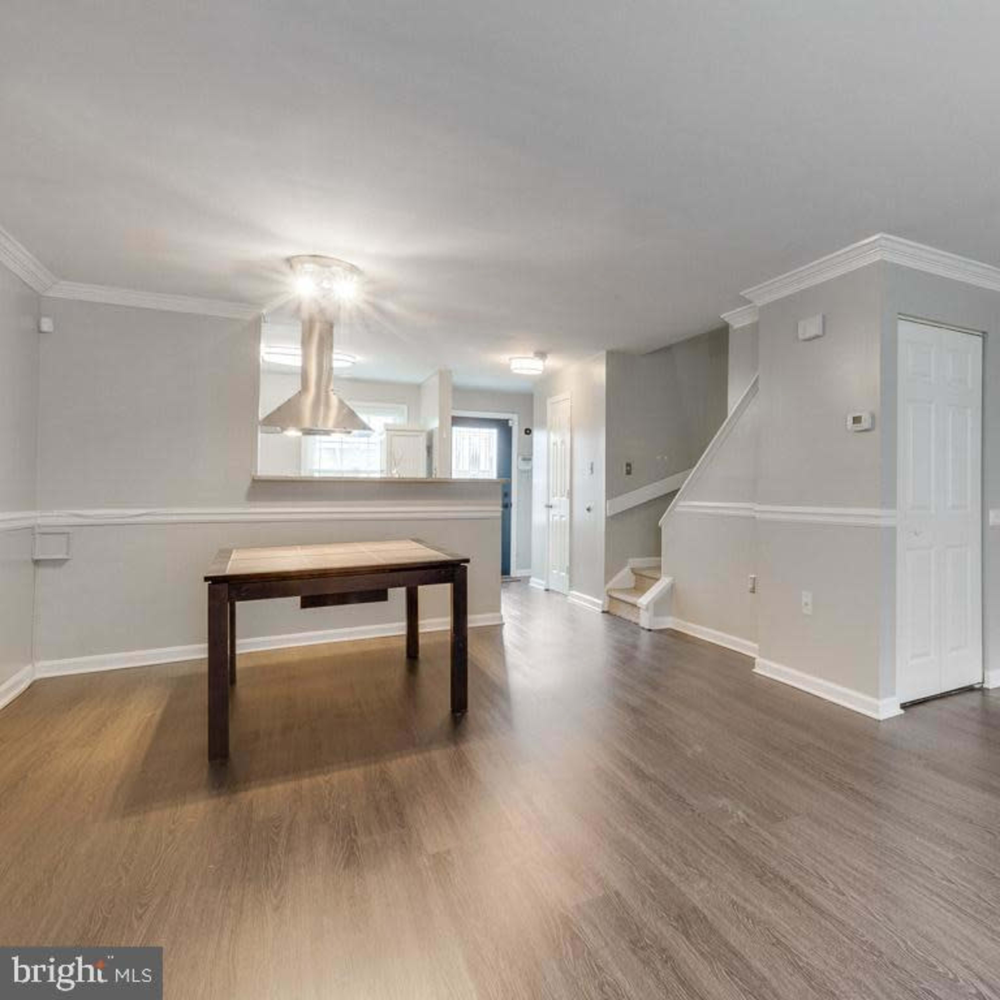 A modern, empty living space featuring a hardwood floor and a table, with a staircase in the background and a kitchen area visible through an open doorway.