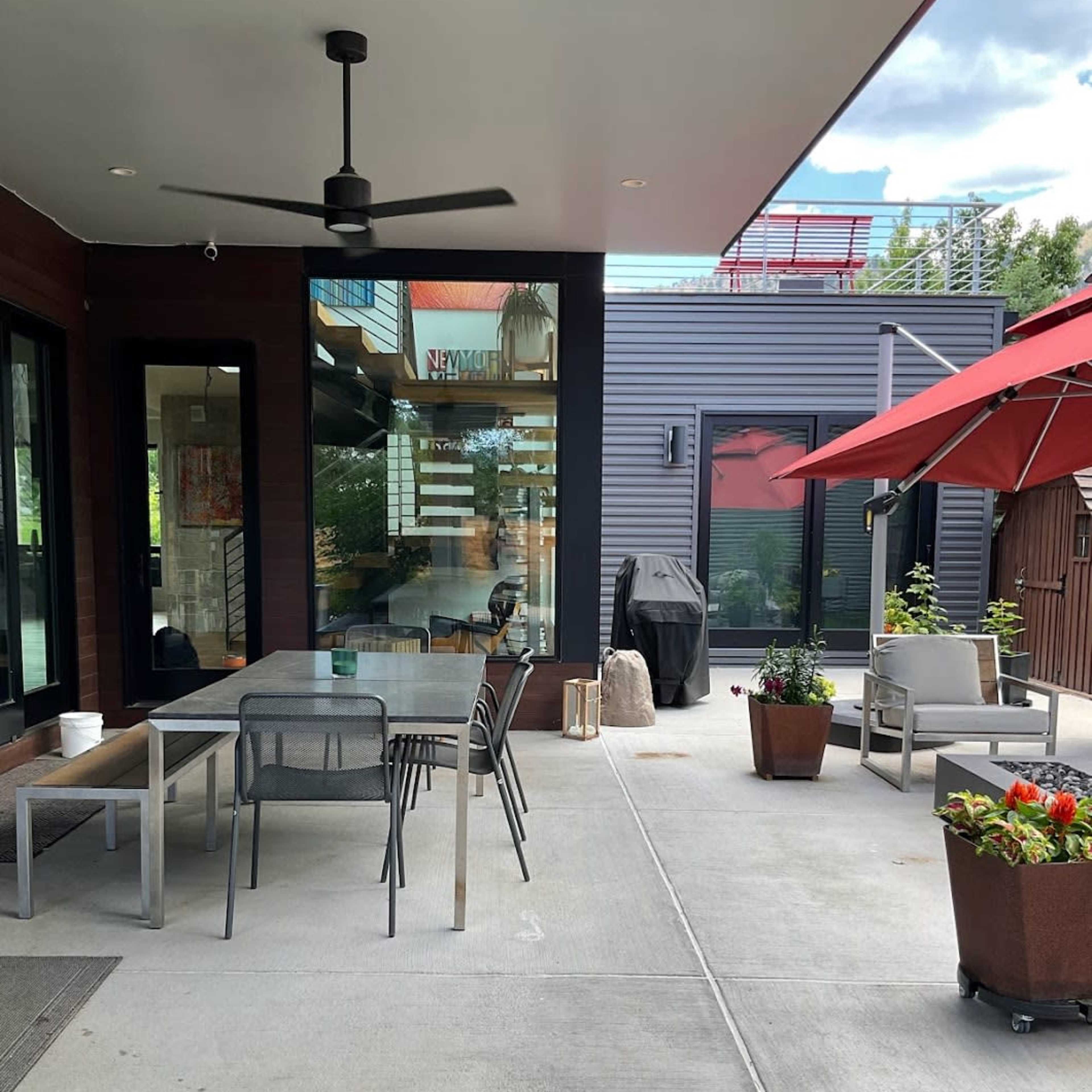 A modern patio area featuring a dining table, chairs, a barbecue grill, and potted plants under a red umbrella.