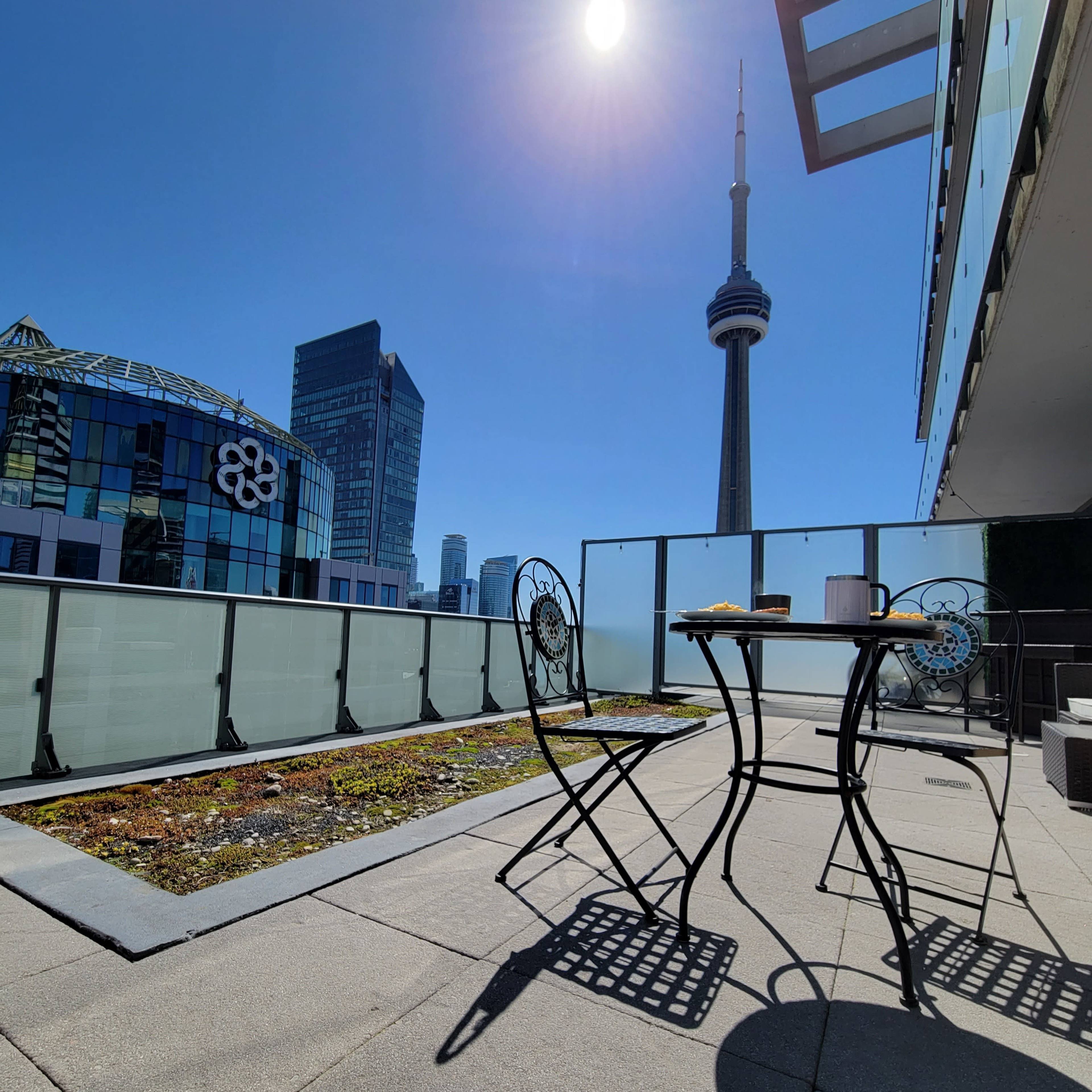A small table with two chairs sits on a rooftop terrace overlooking the CN Tower and nearby buildings under a clear blue sky.