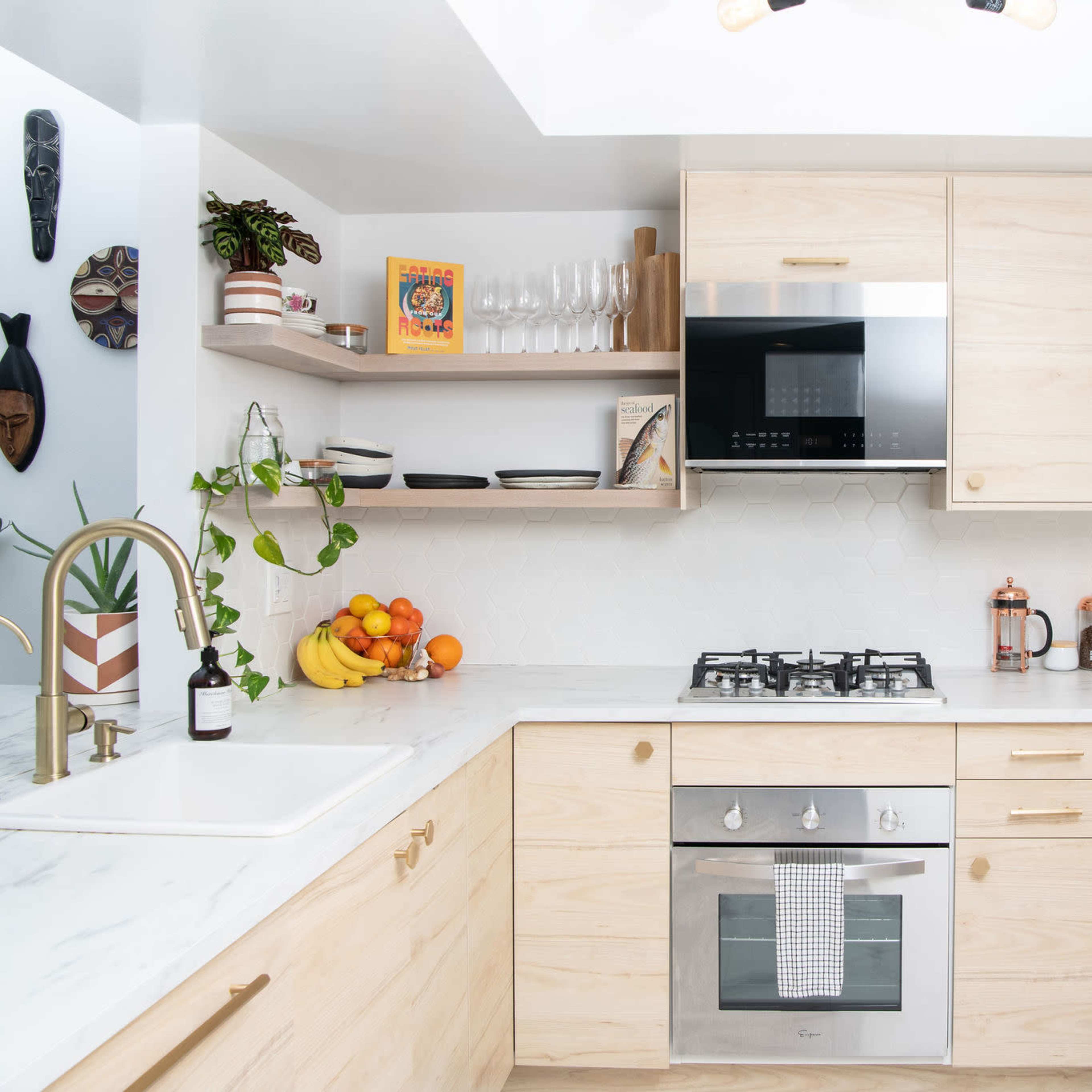 A modern kitchen features light wood cabinetry, a white marble countertop, and a gas stove, with fruit on the counter and houseplants hanging in macramé planters.