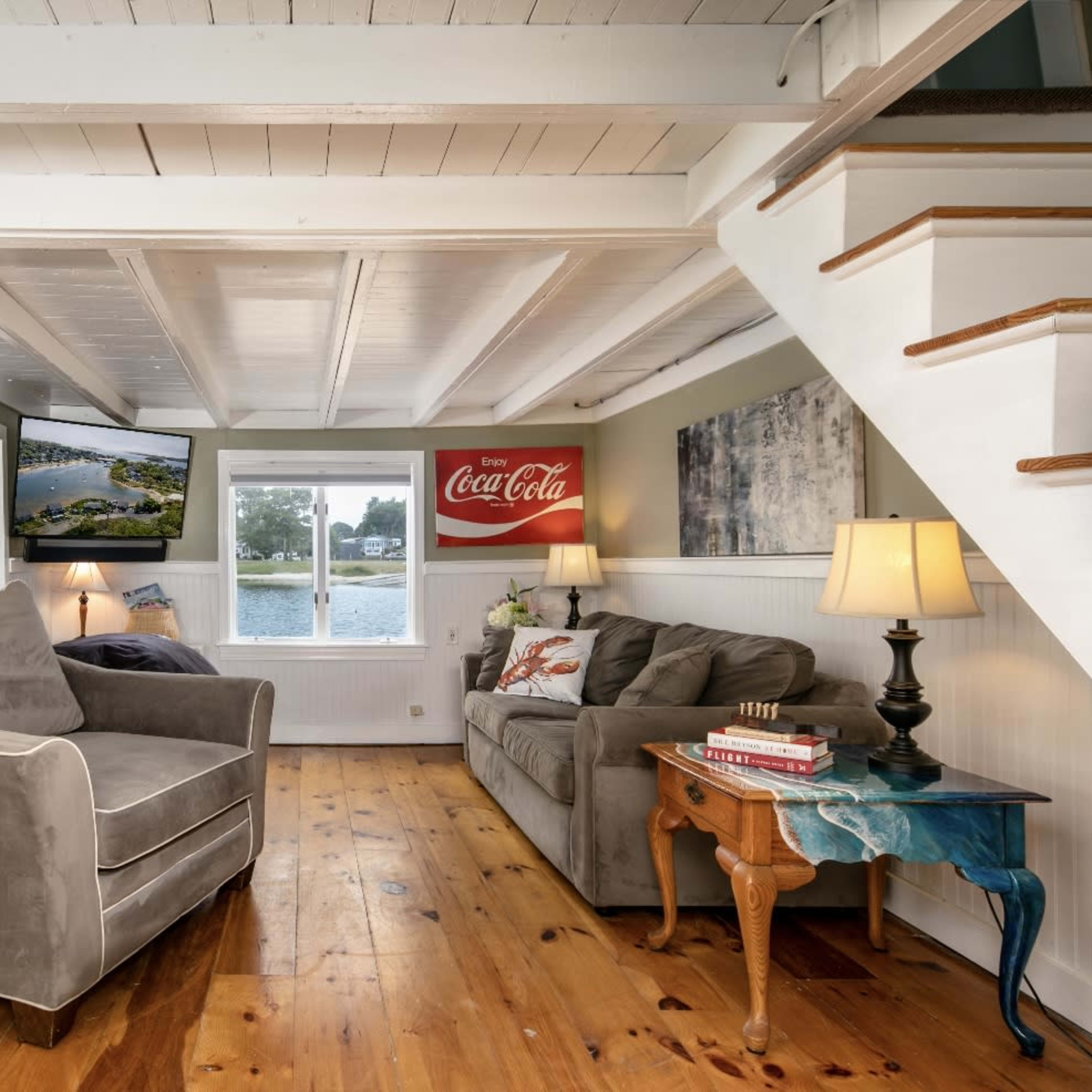 A cozy living room with a gray armchair and couch, a wooden floor, wall-mounted TV, and a staircase leading to an upper level.