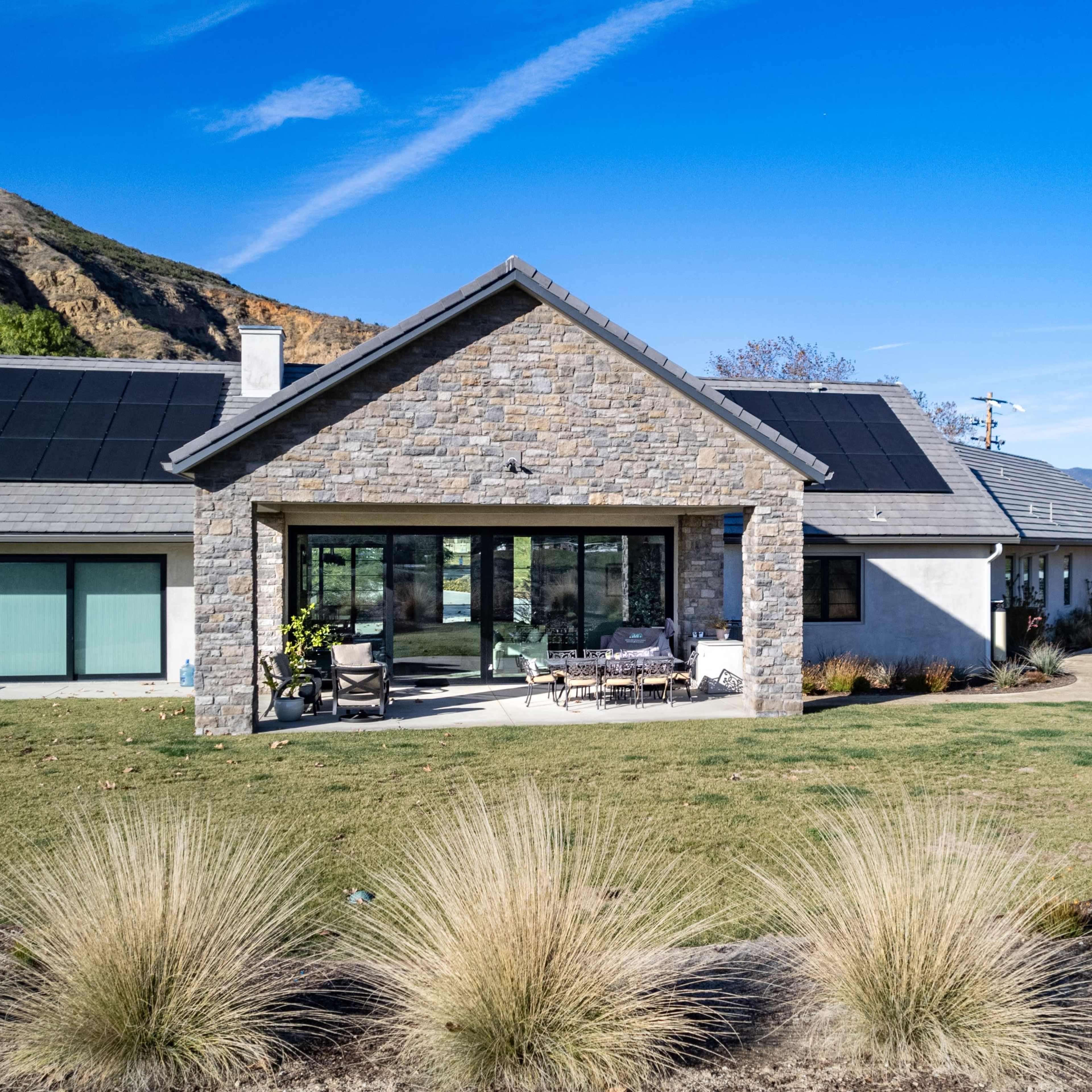 A modern house with solar panels on the roof is surrounded by landscaped gardens and mountains in the background.