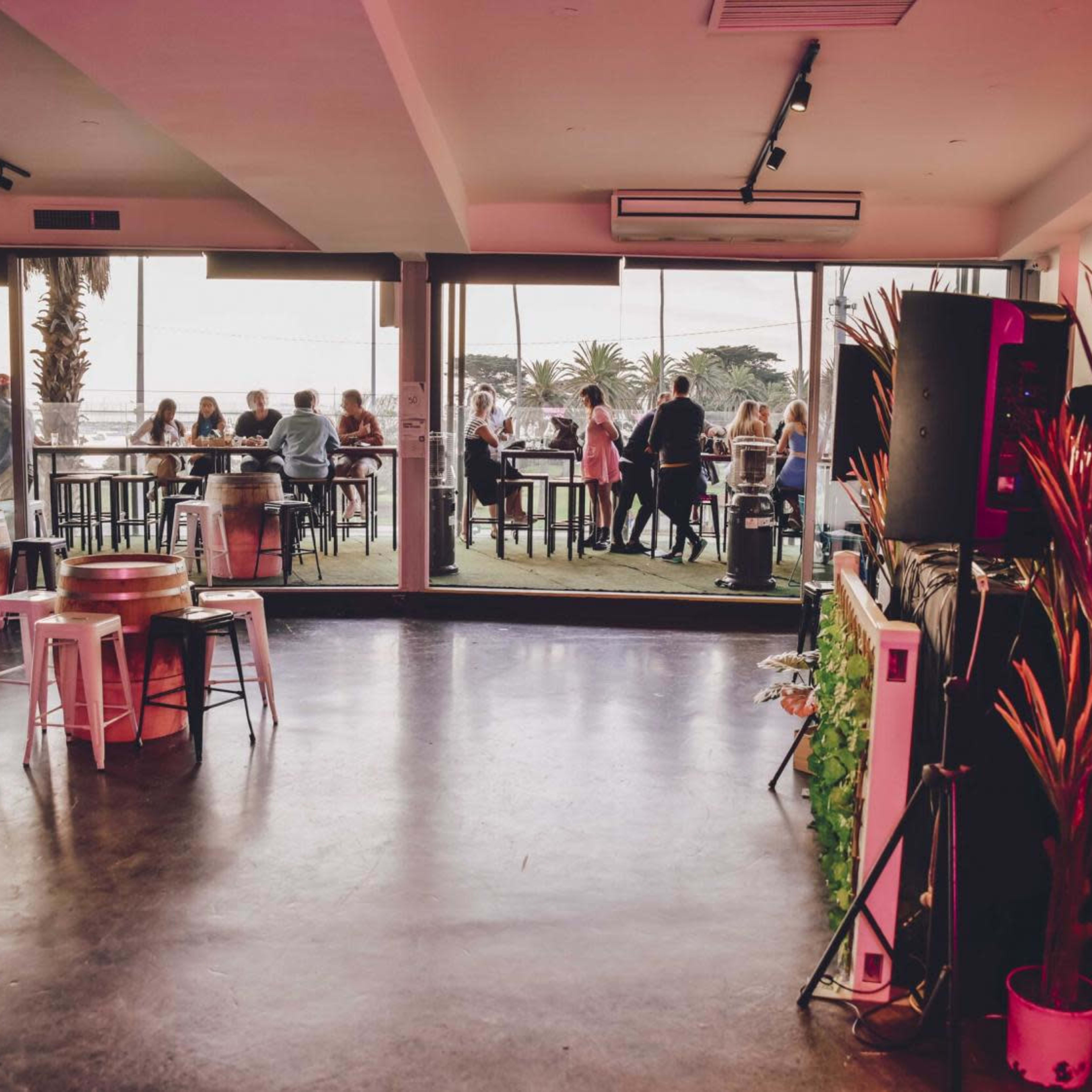 The interior of a venue features seating by large windows overlooking a social gathering on a patio.
