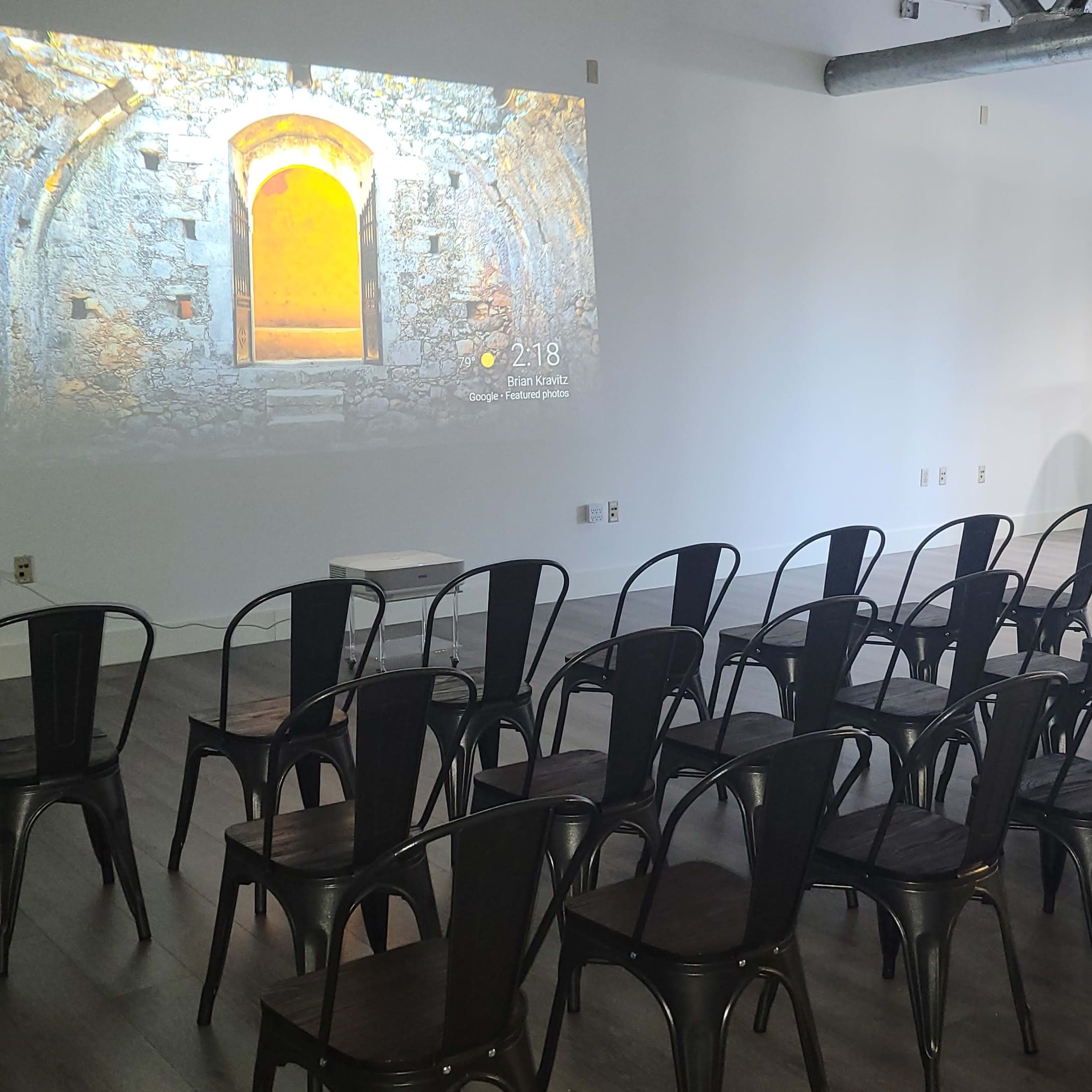 A room with rows of black metal chairs facing a projected image of a stone wall with a doorway.