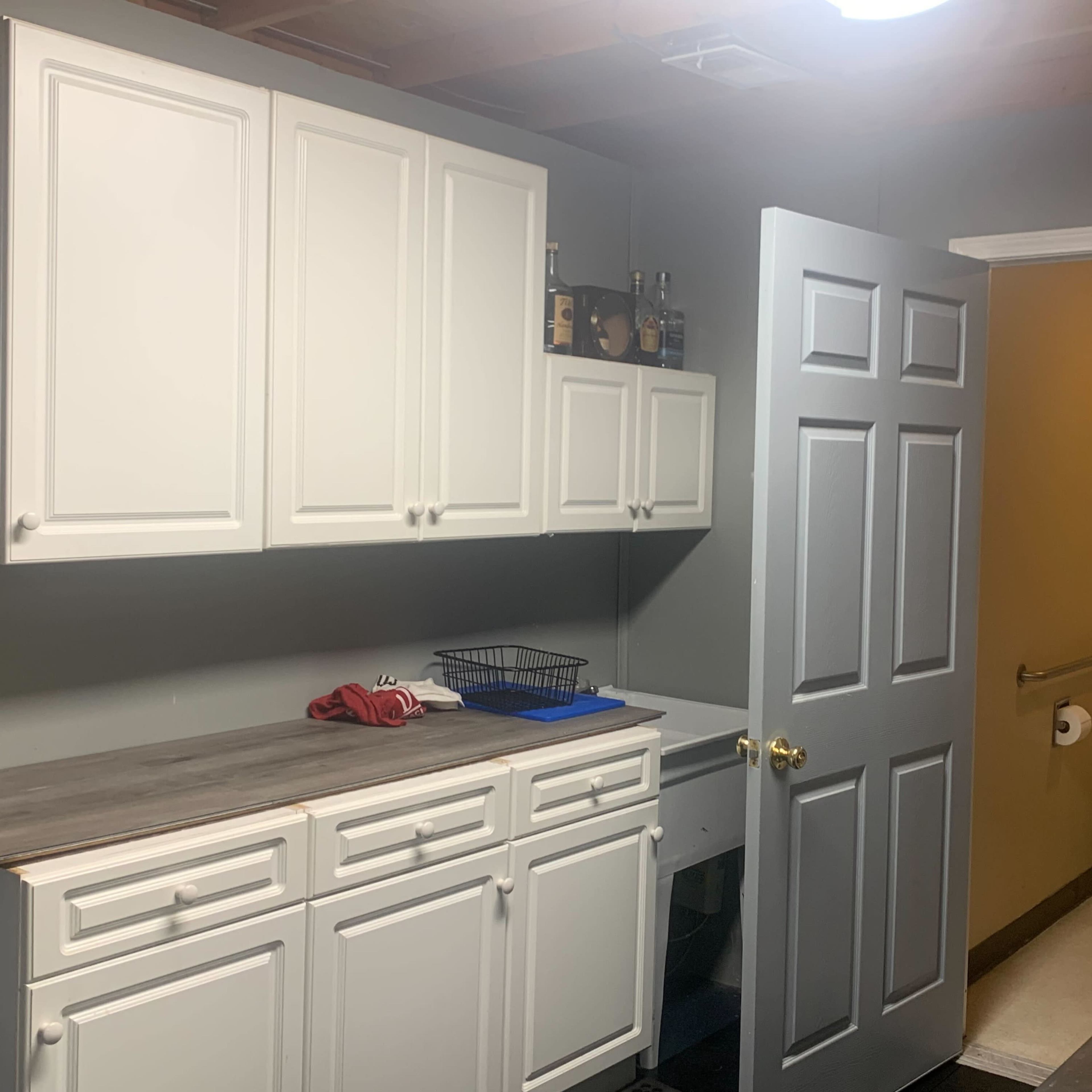 A laundry room with white cabinets, a countertop, and an open door leading to a bathroom.