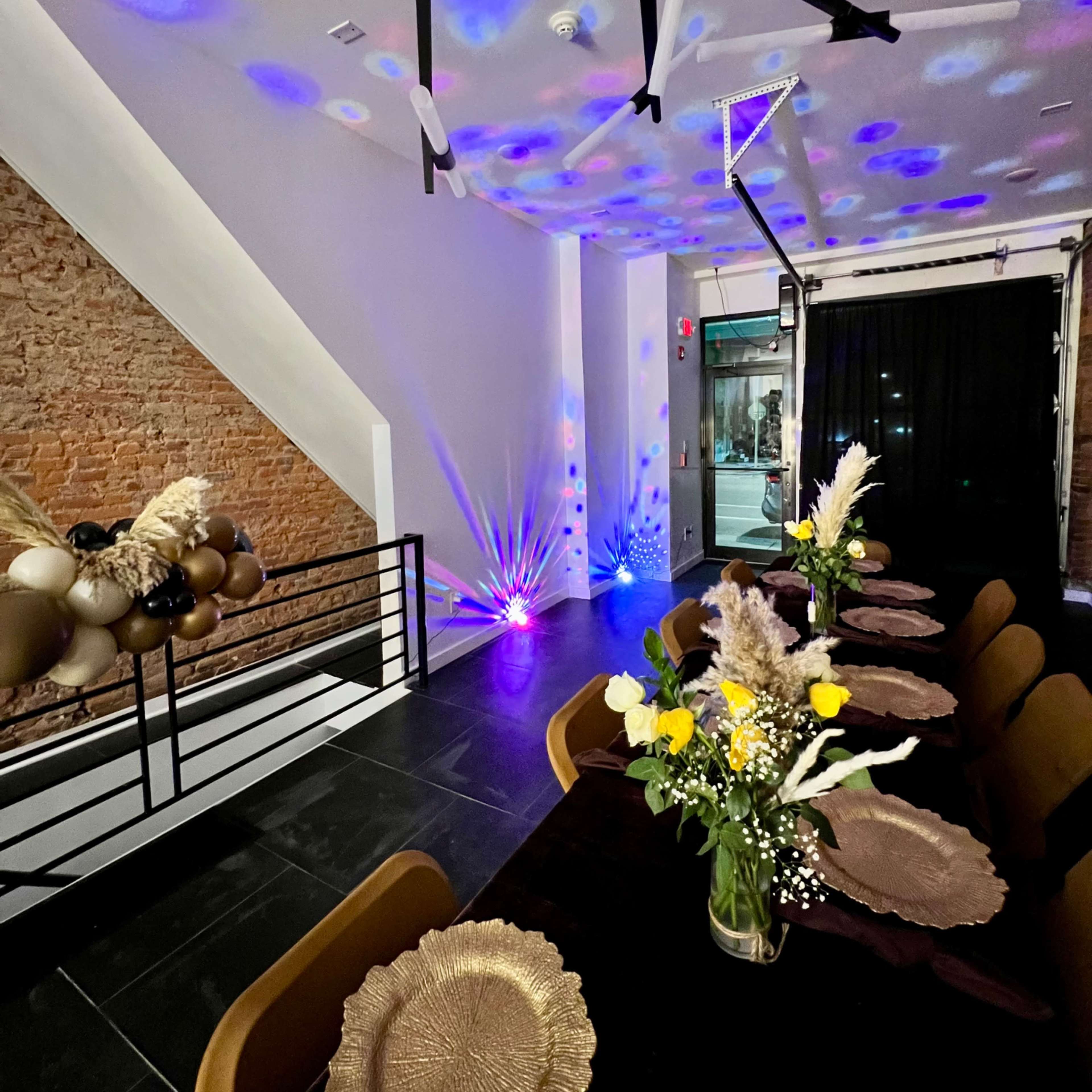 A modern event space with a long dining table set with decorative plates, illuminated by colorful lights on the ceiling and featuring a brick wall in the background.