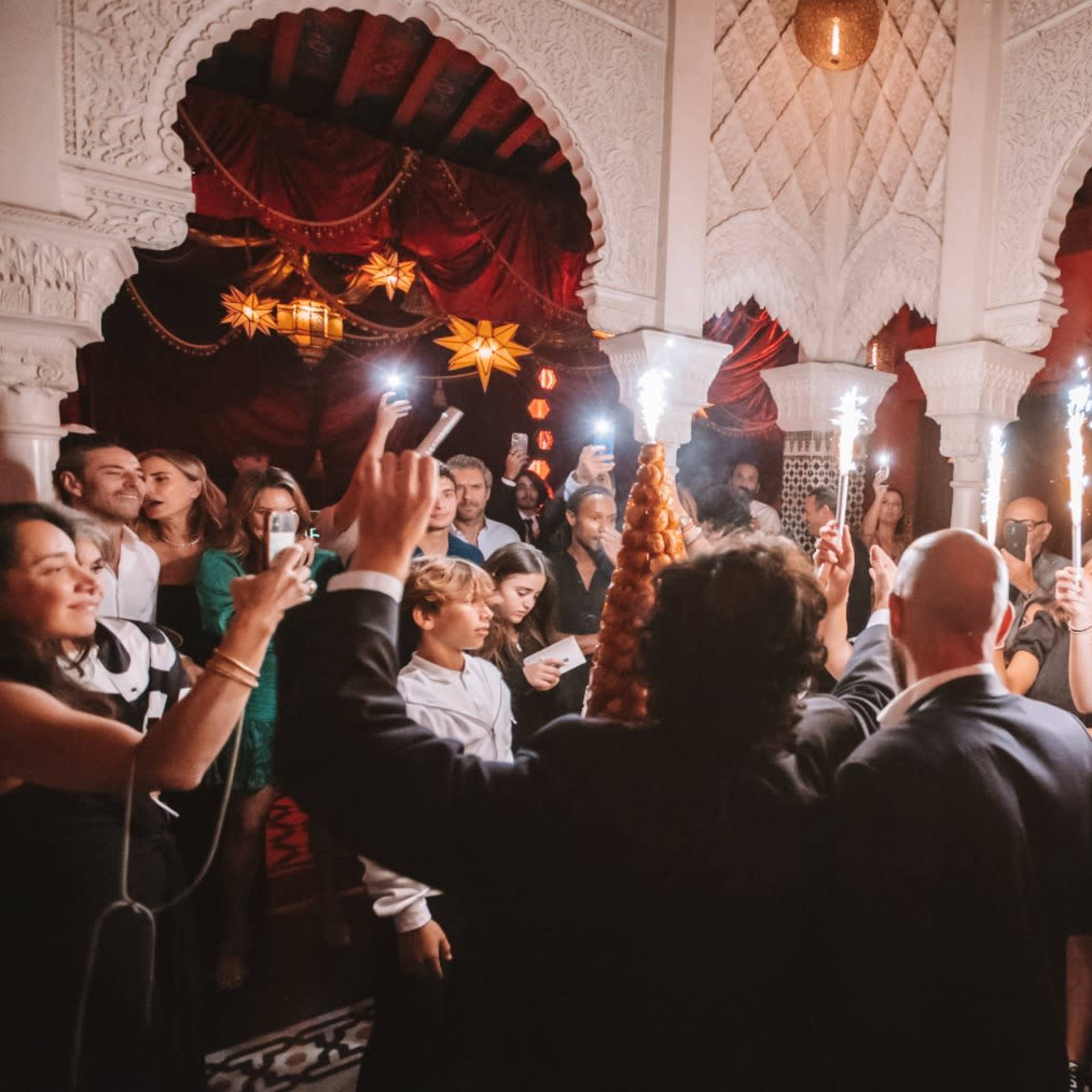 A group of people gather in an ornate room, raising their phones and lights while celebrating an event.
