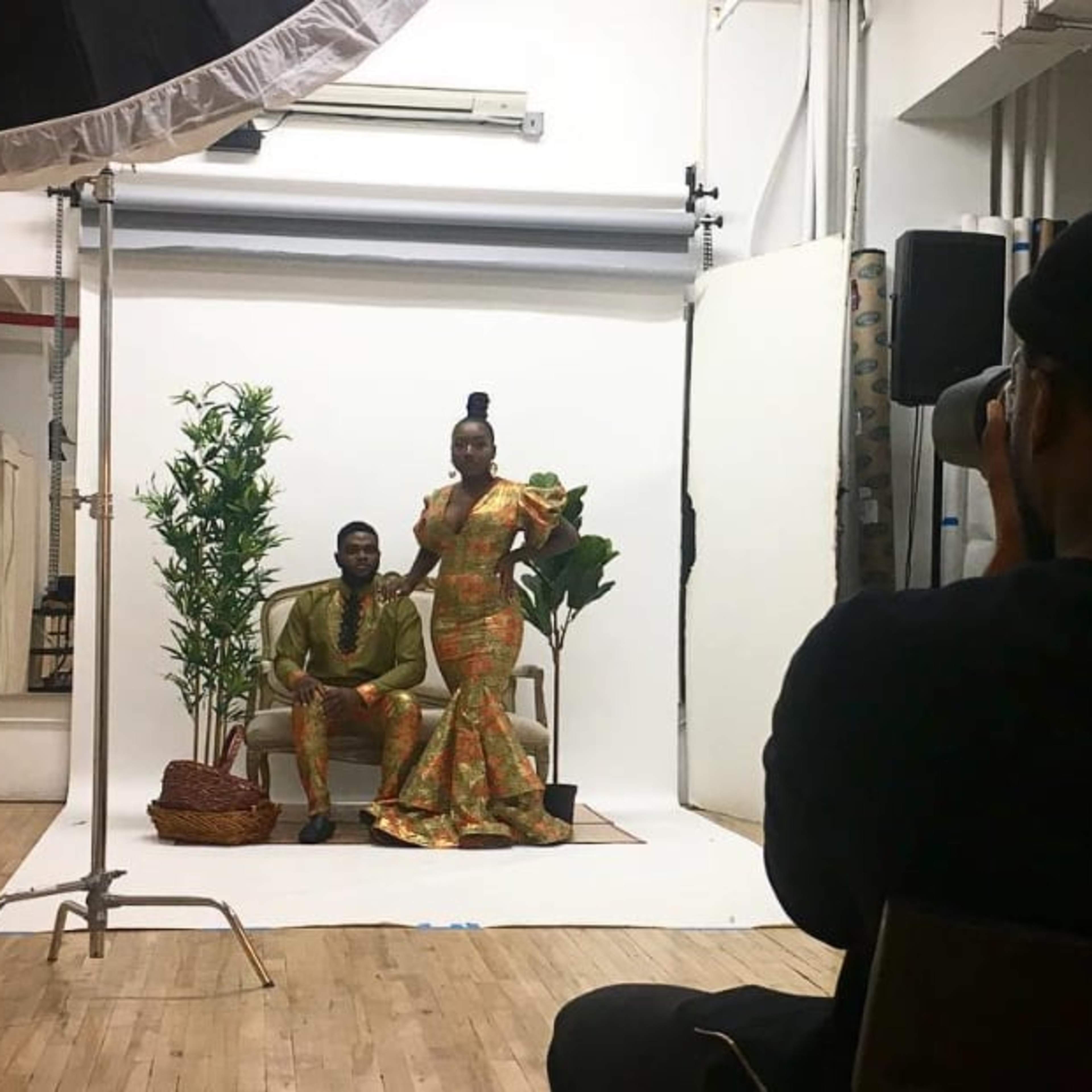 A photographer captures a couple posing on a couch in a studio setting with plants and a backdrop.