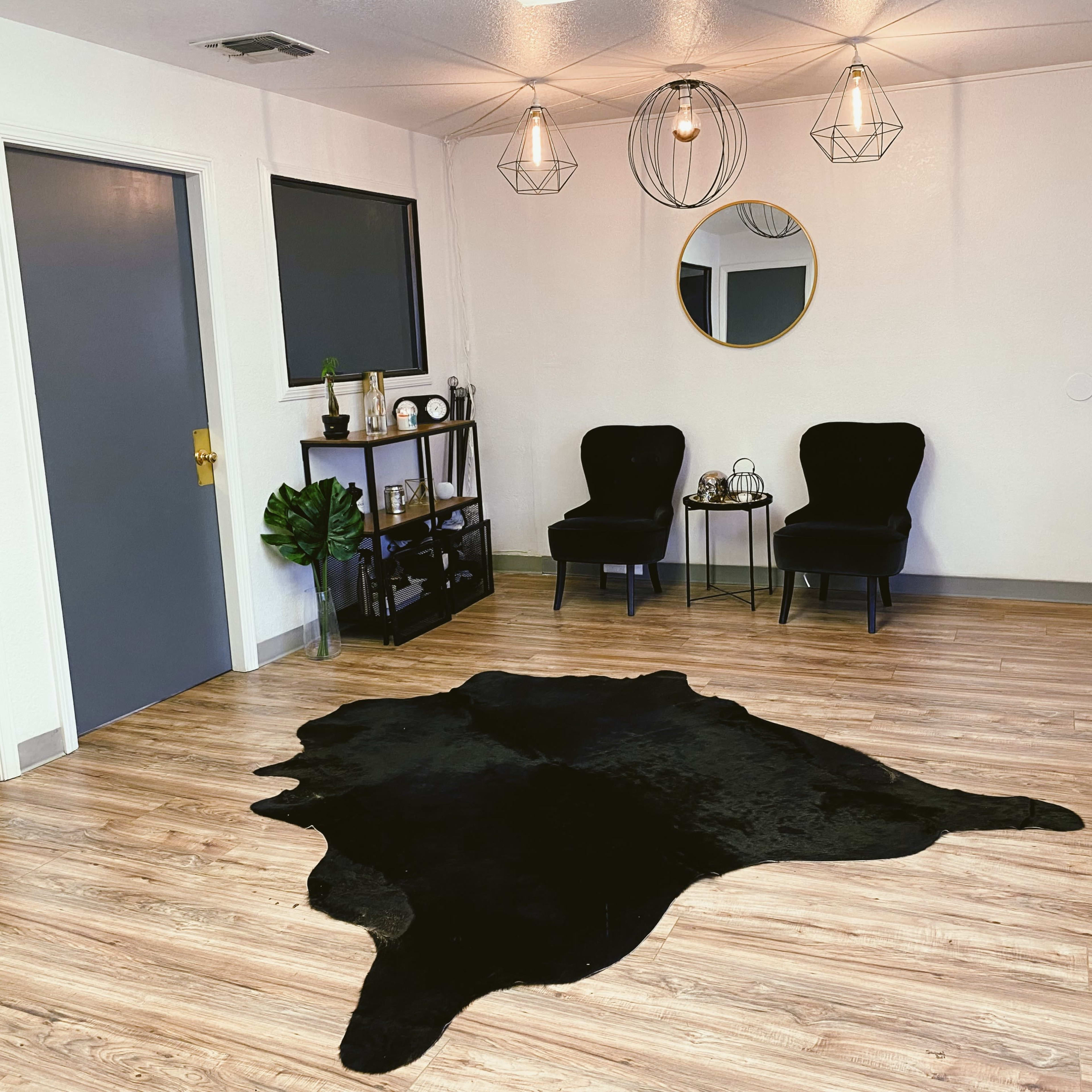 A minimalist room with two black chairs, a round mirror, and a black rug on wooden flooring, accompanied by a small shelving unit and potted plants.