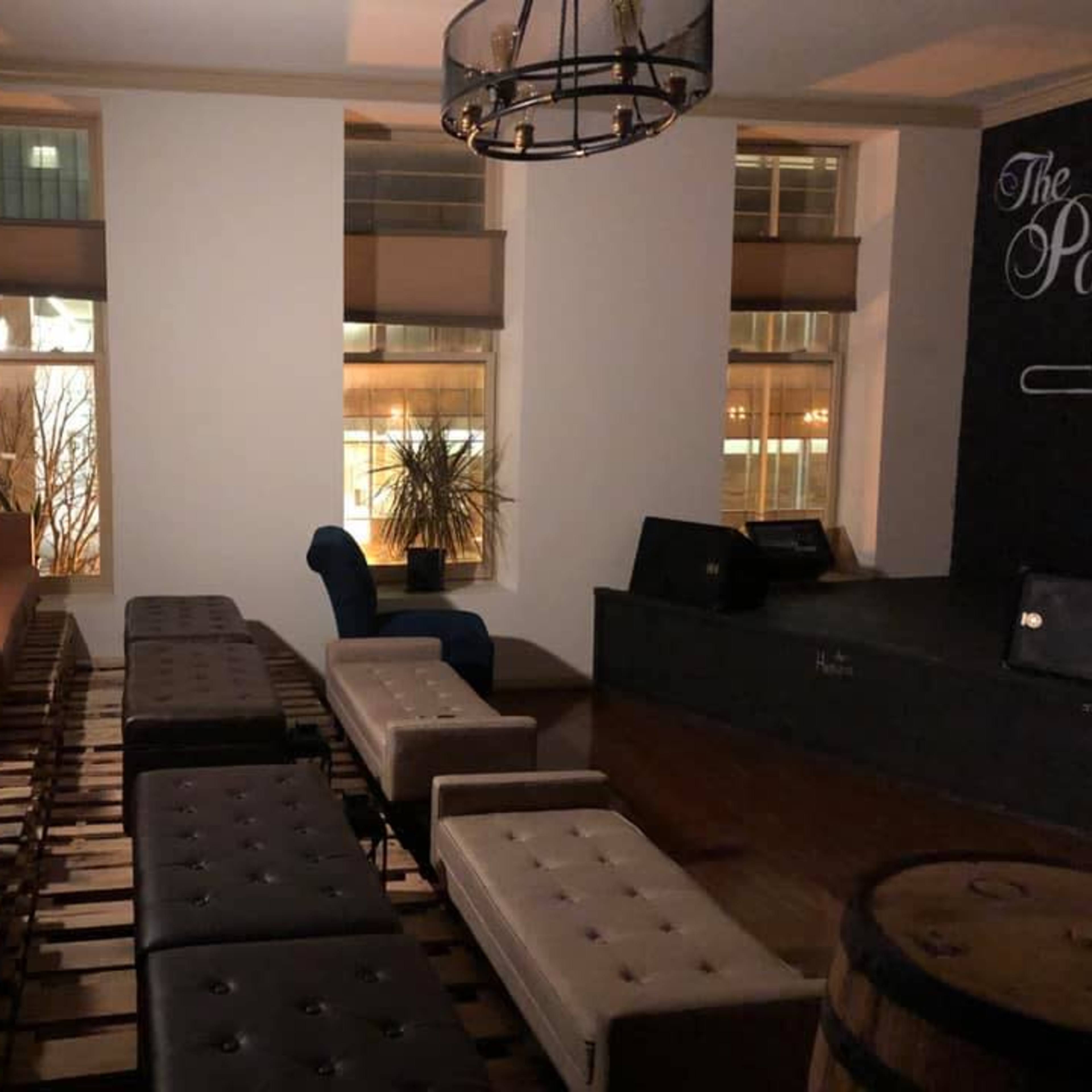 A cozy lounge area with a mix of seating, including a brown leather couch, white ottomans, and a blue chair, illuminated by soft lighting from windows.