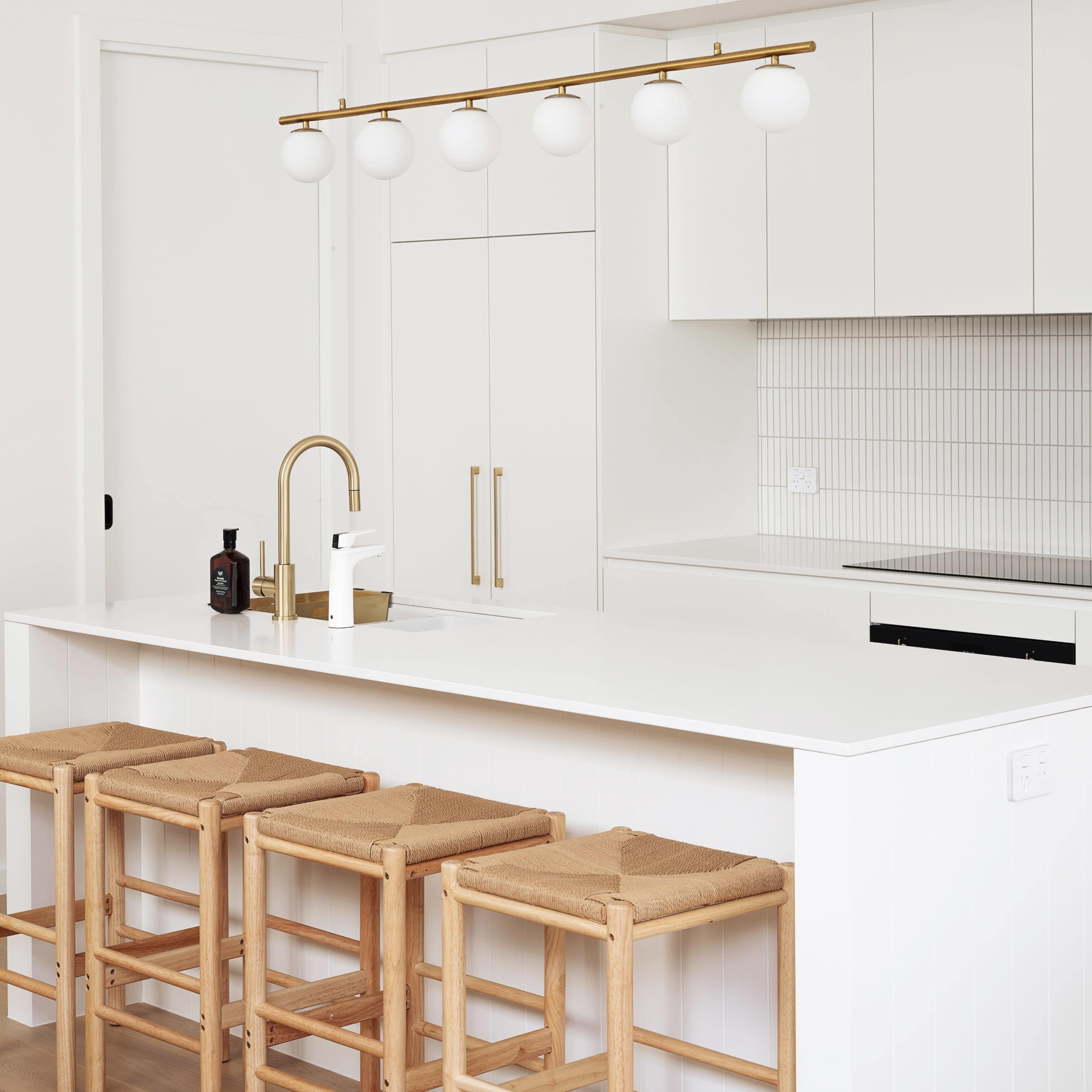 A modern kitchen features a white countertop with three wooden stools, a brass faucet, and a light fixture with five spherical lights hanging above.