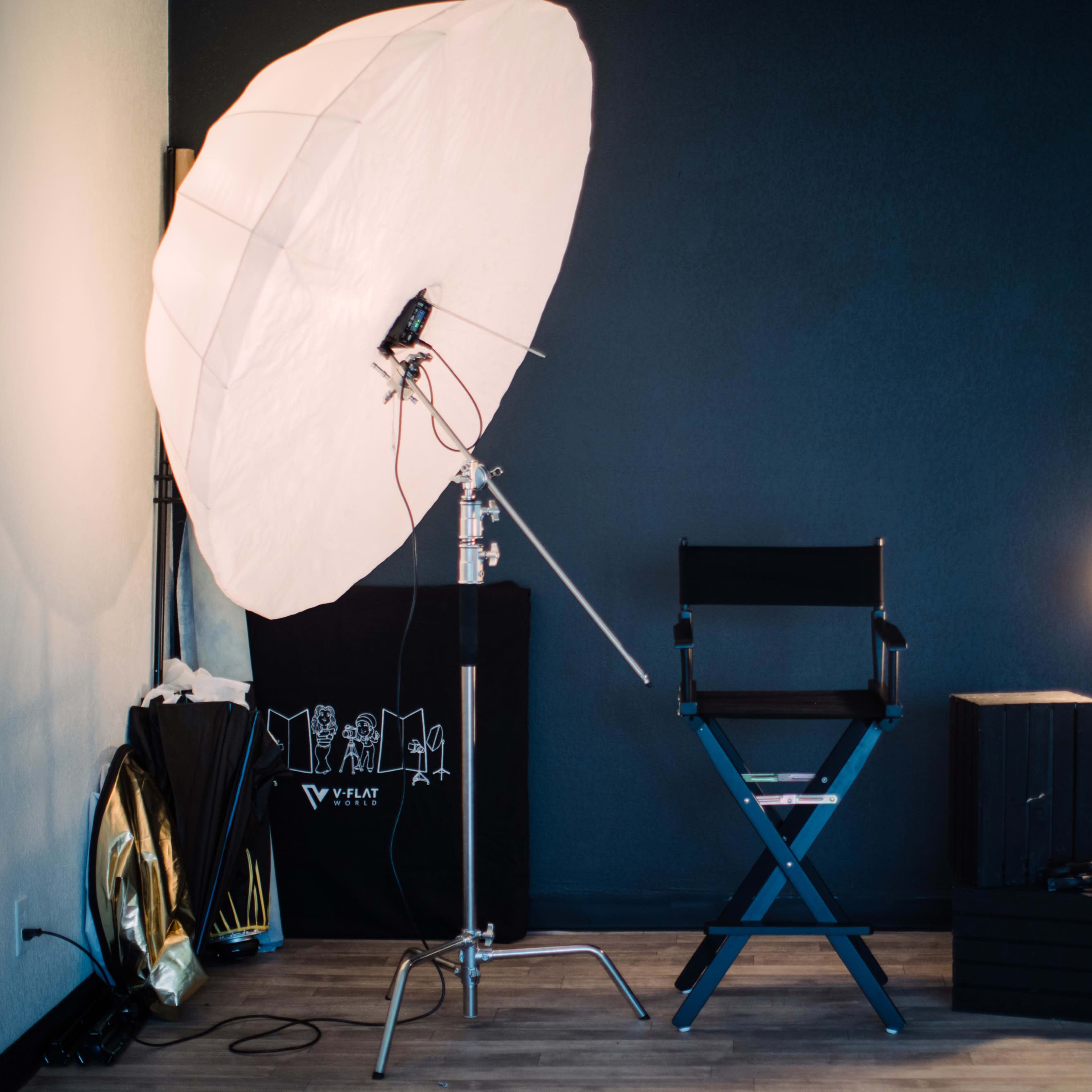A photography studio setup features a large soft umbrella light, a director's chair, and additional lighting equipment against a dark wall.