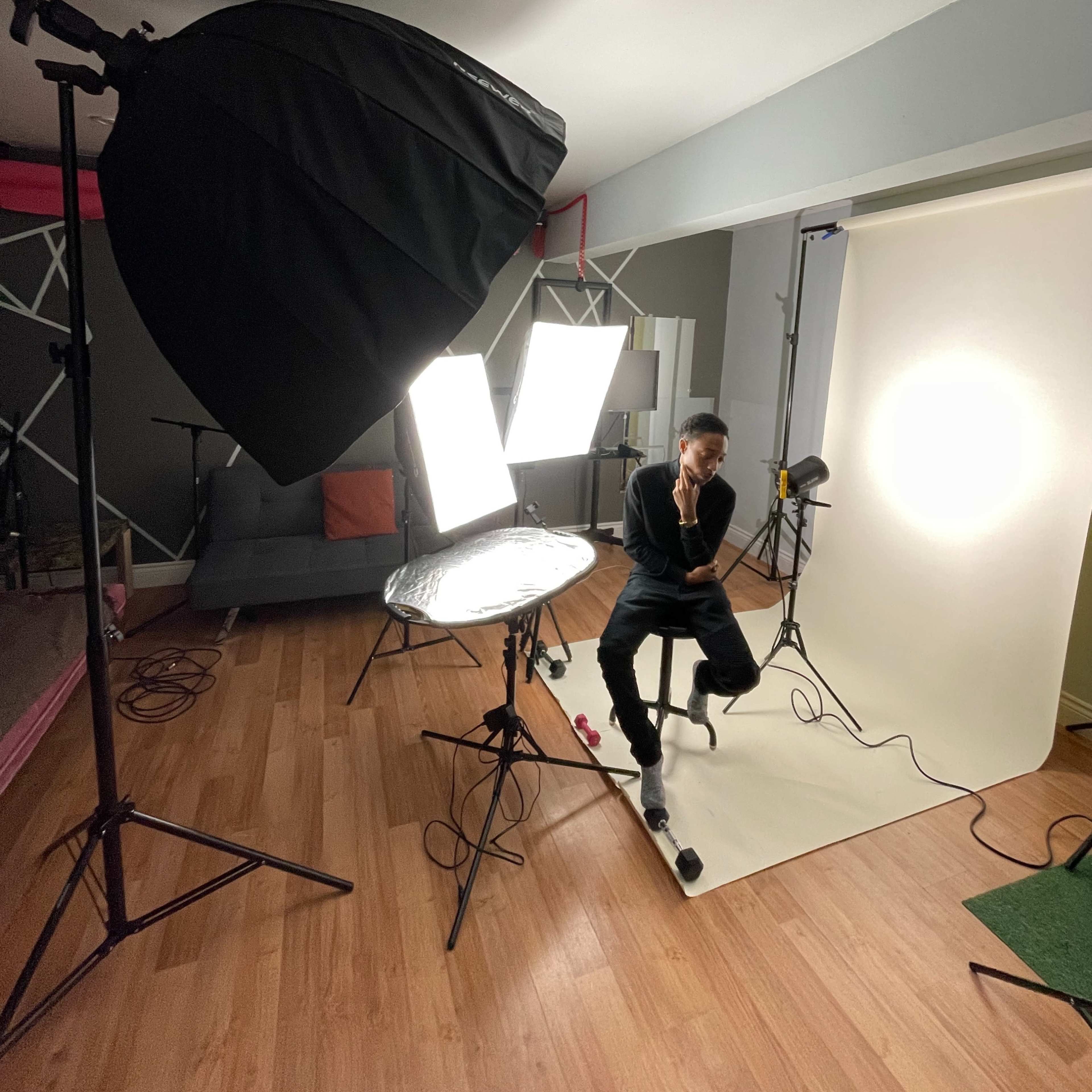 A person sits on a stool in a photography studio with several light sources and a backdrop set up for a photoshoot.