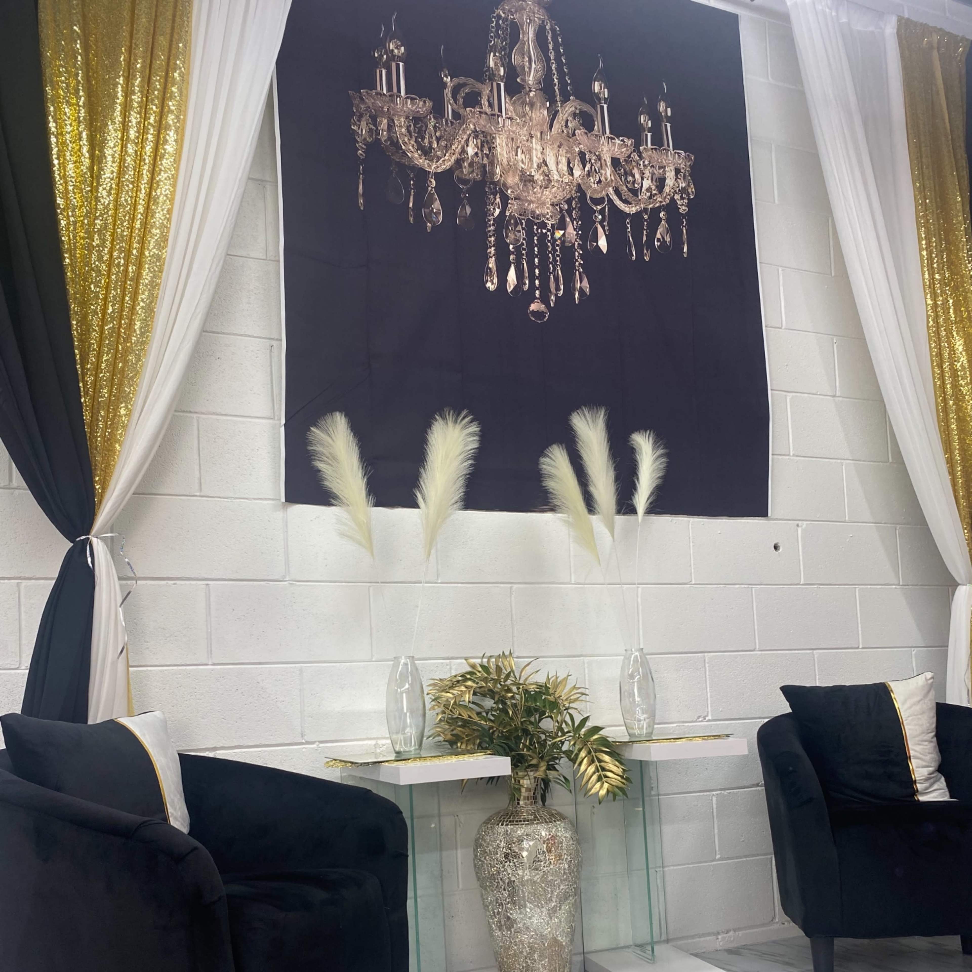 A stylish interior showcases two black chairs opposite a glass table adorned with a decorative vase and tall, feathery arrangements, framed by black and gold drapery.