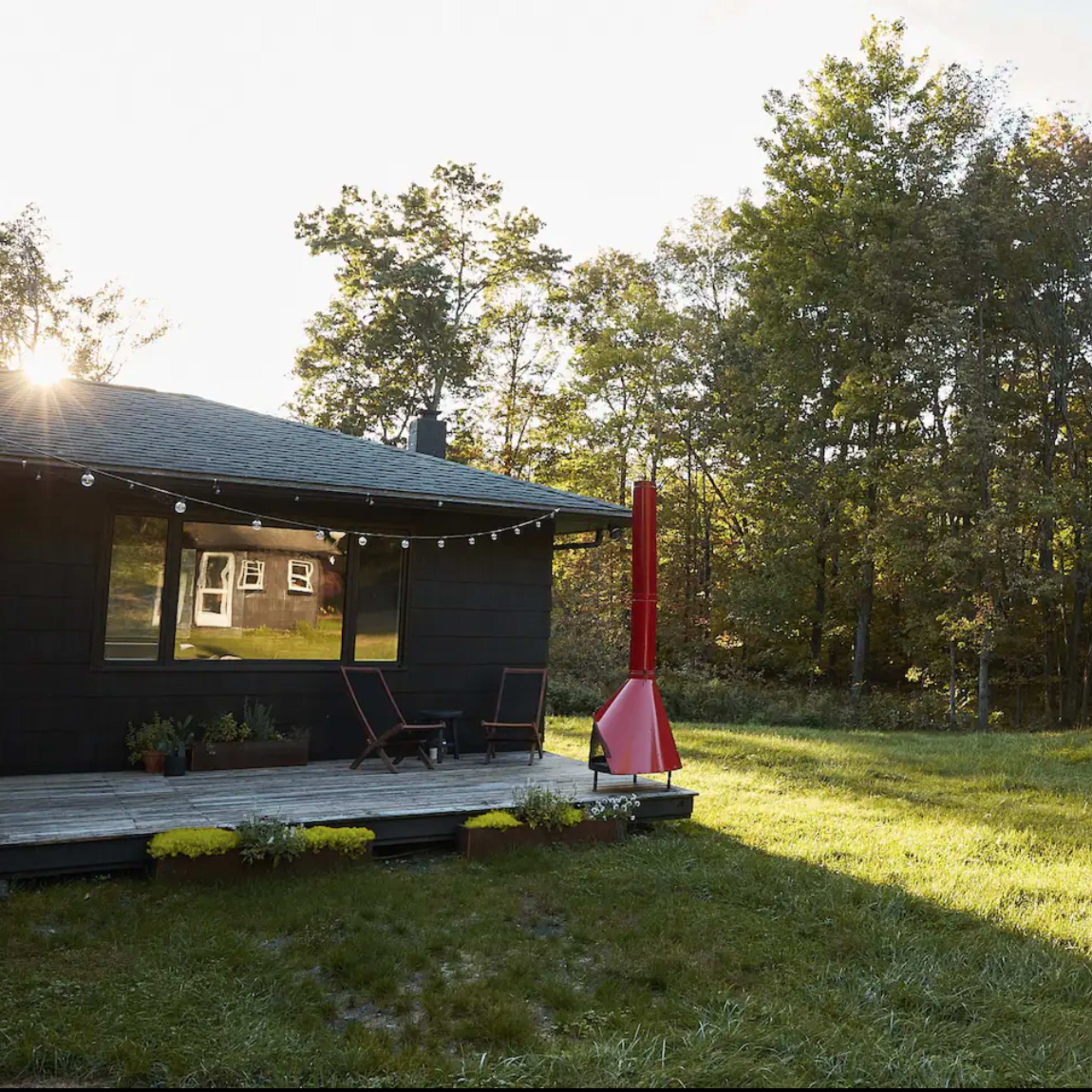 A modern cabin with large windows and a red chimney stands on a wooden deck in a grassy area surrounded by trees.