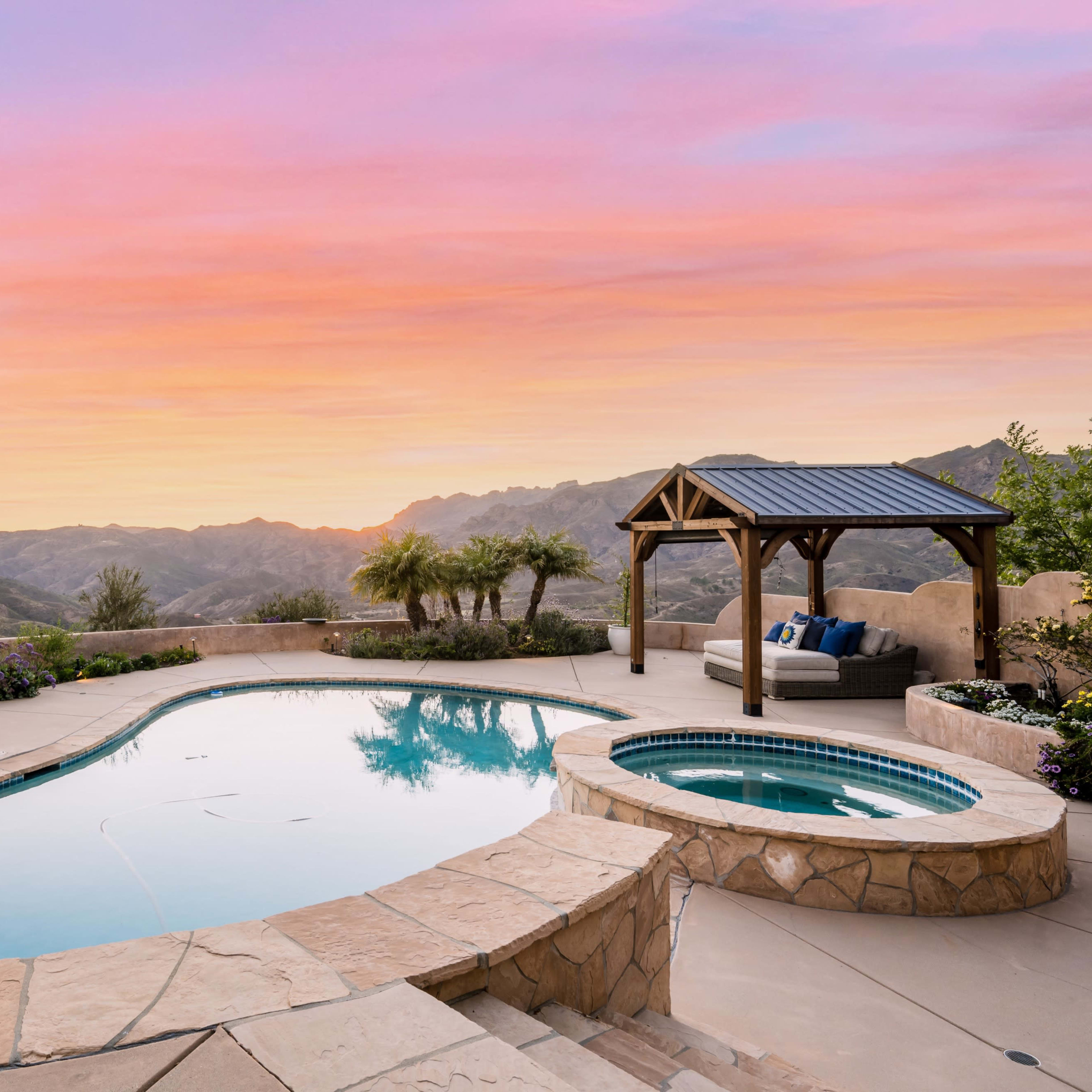 A stone patio surrounds a swimming pool with a spa, overlooking rolling hills under a colorful sunset sky.