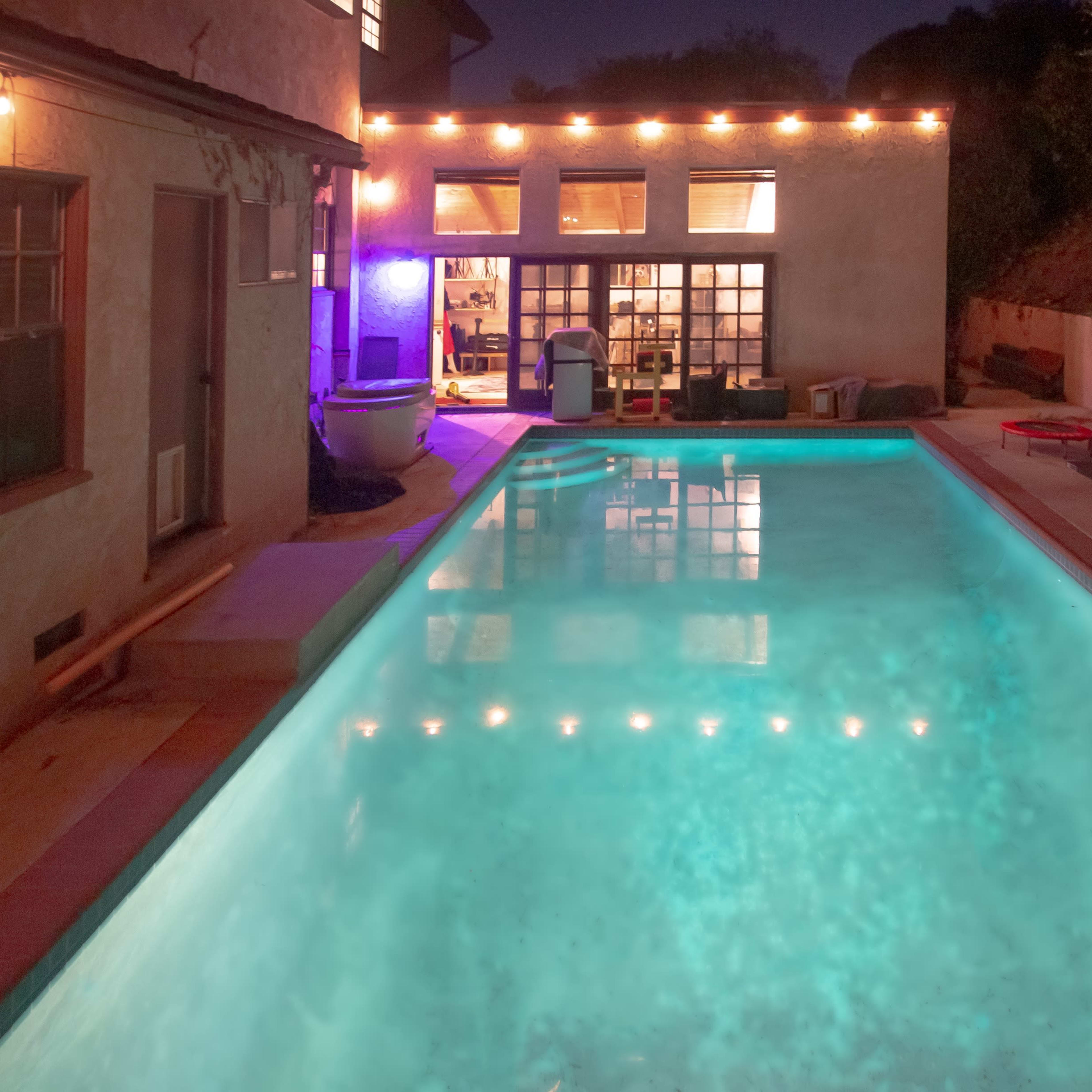 A rectangular swimming pool illuminated by string lights is situated beside a house with large windows showing light from inside.