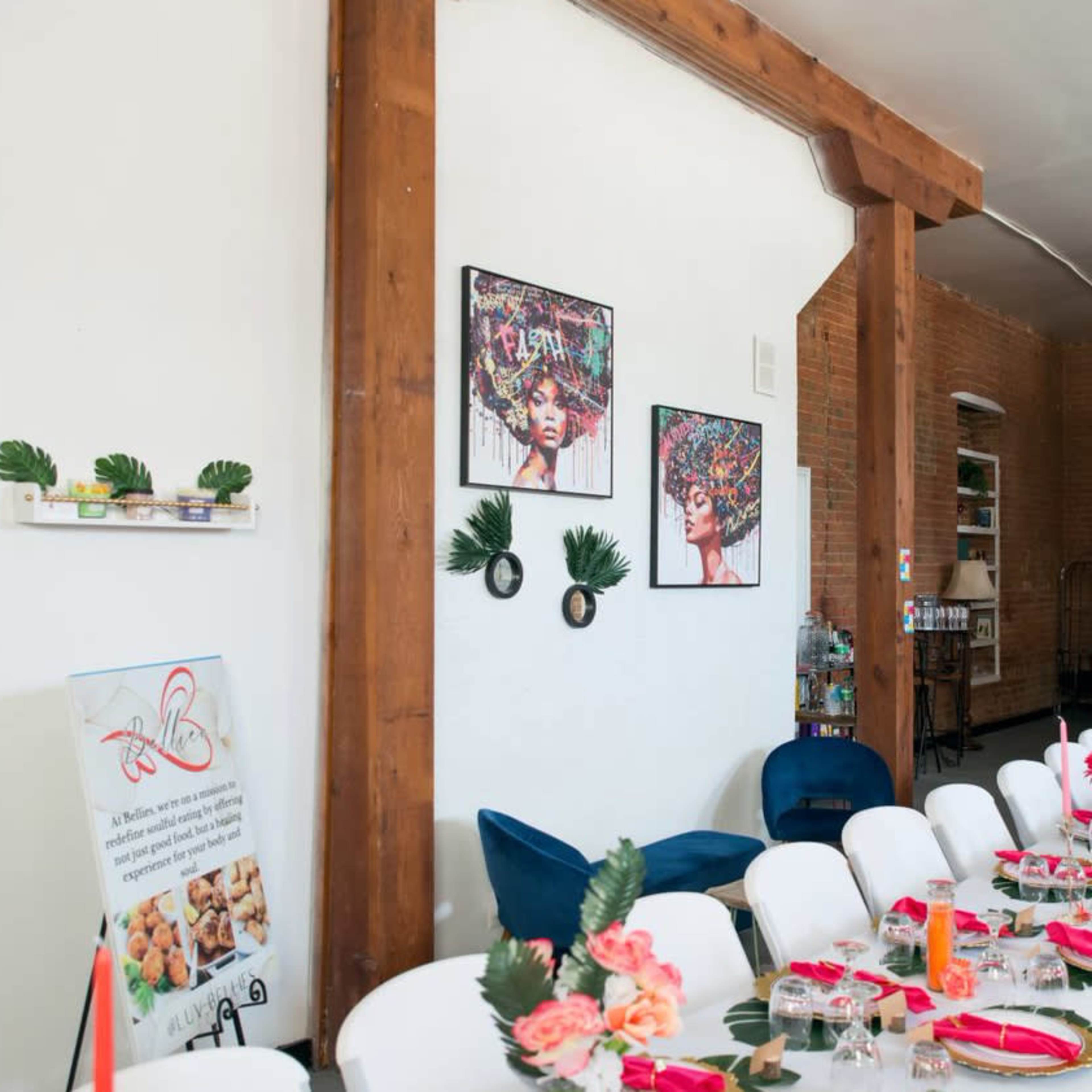 A long dining table is set with pink napkins and flowers, surrounded by chairs in a room featuring exposed wooden beams and colorful artwork on the walls.