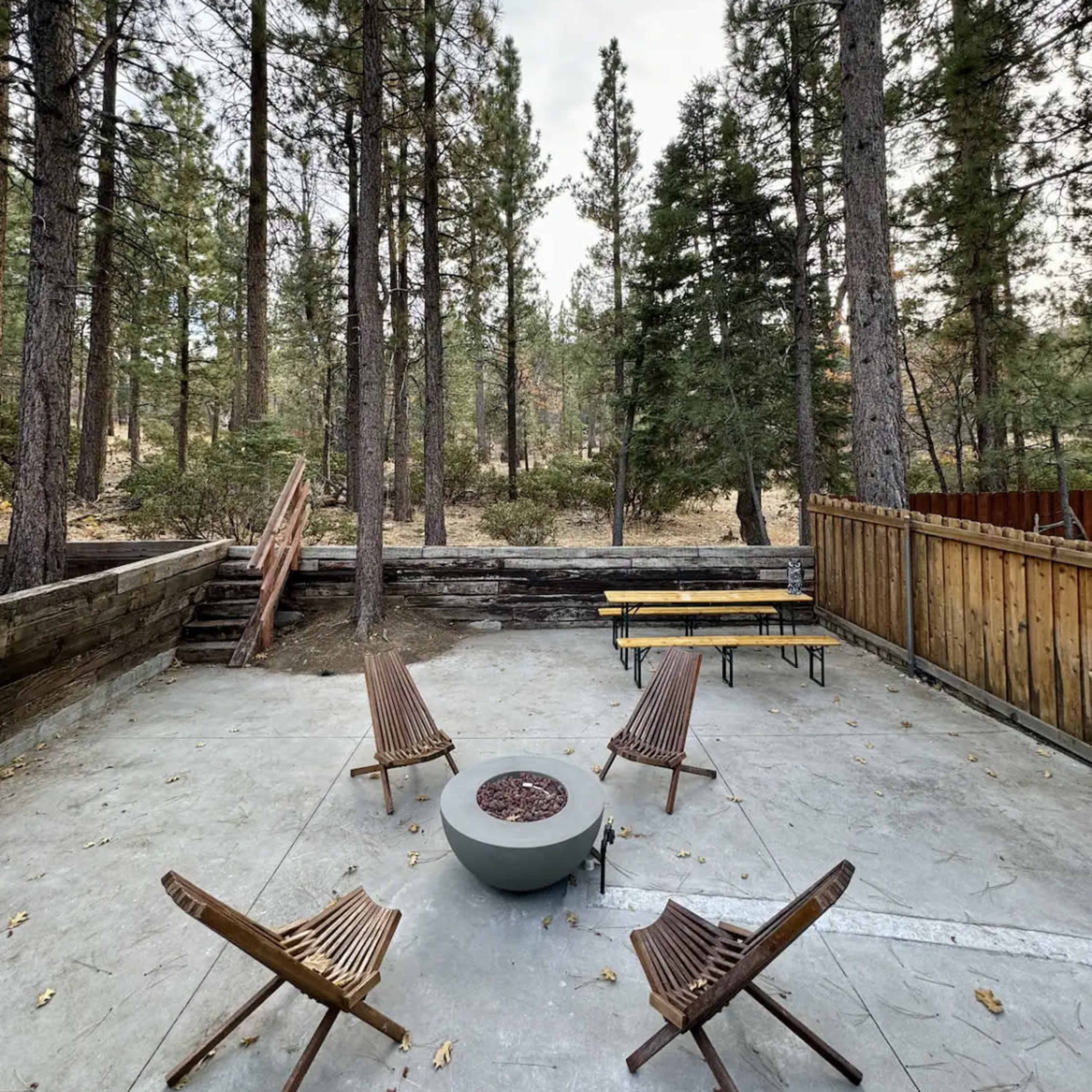 A concrete patio features four wooden chairs arranged around a central fire pit, surrounded by tall pine trees.
