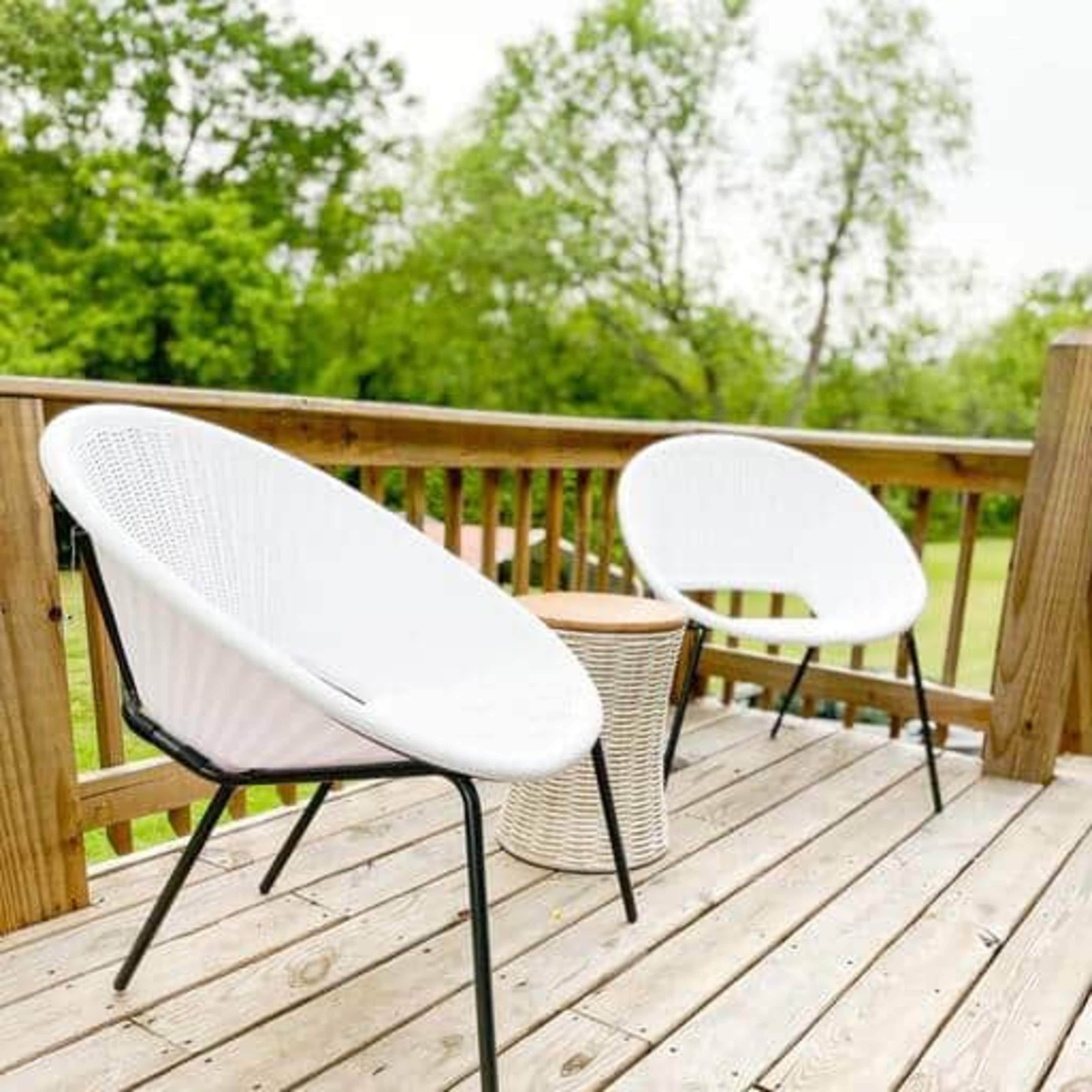 Two white chairs and a small table are placed on a wooden deck surrounded by greenery.