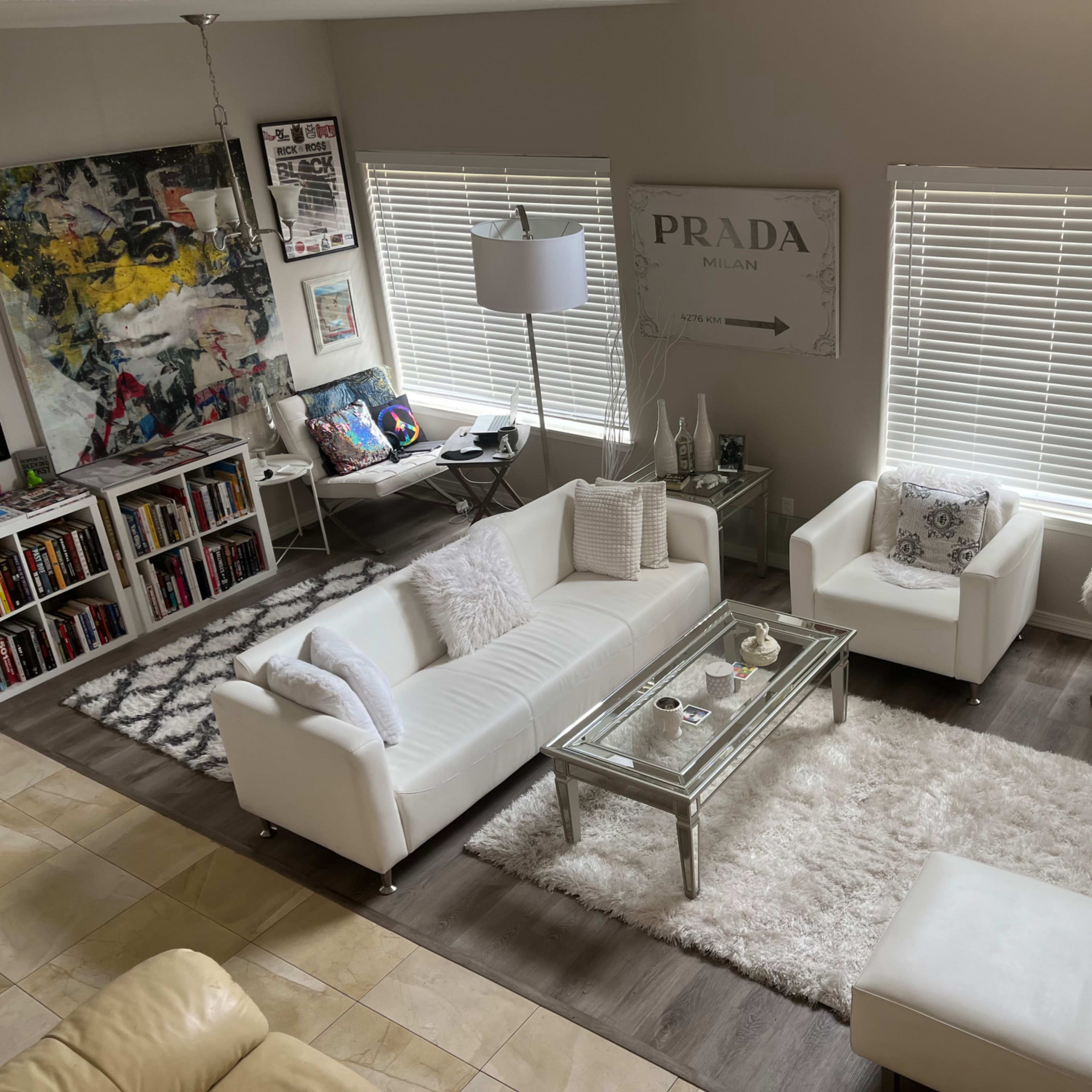 A modern living room with white furniture, a glass coffee table, and large windows with blinds, featuring a mix of decorative items and artworks on the walls.