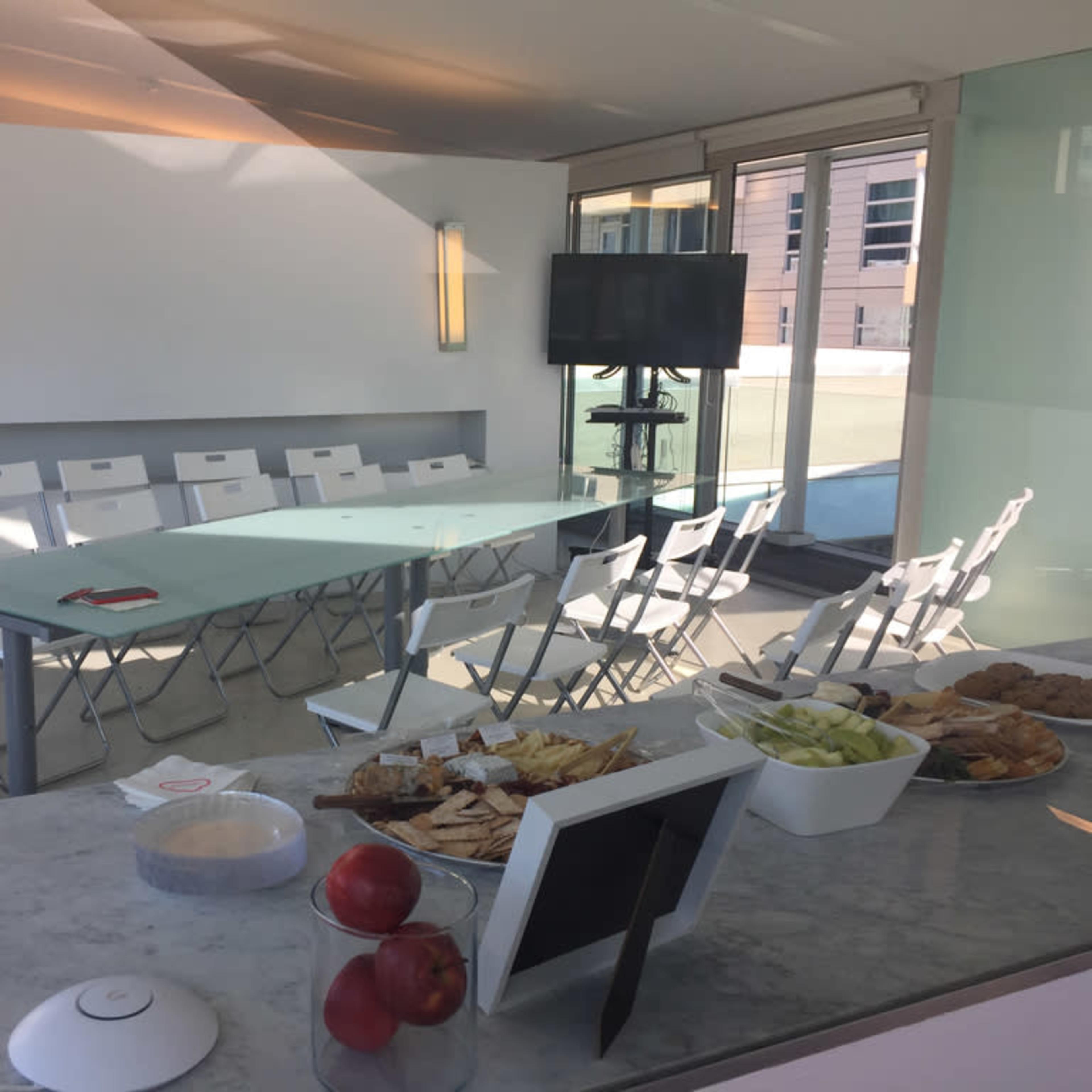 A modern conference room features a long table surrounded by white chairs, with a spread of snacks and drinks on a nearby counter.