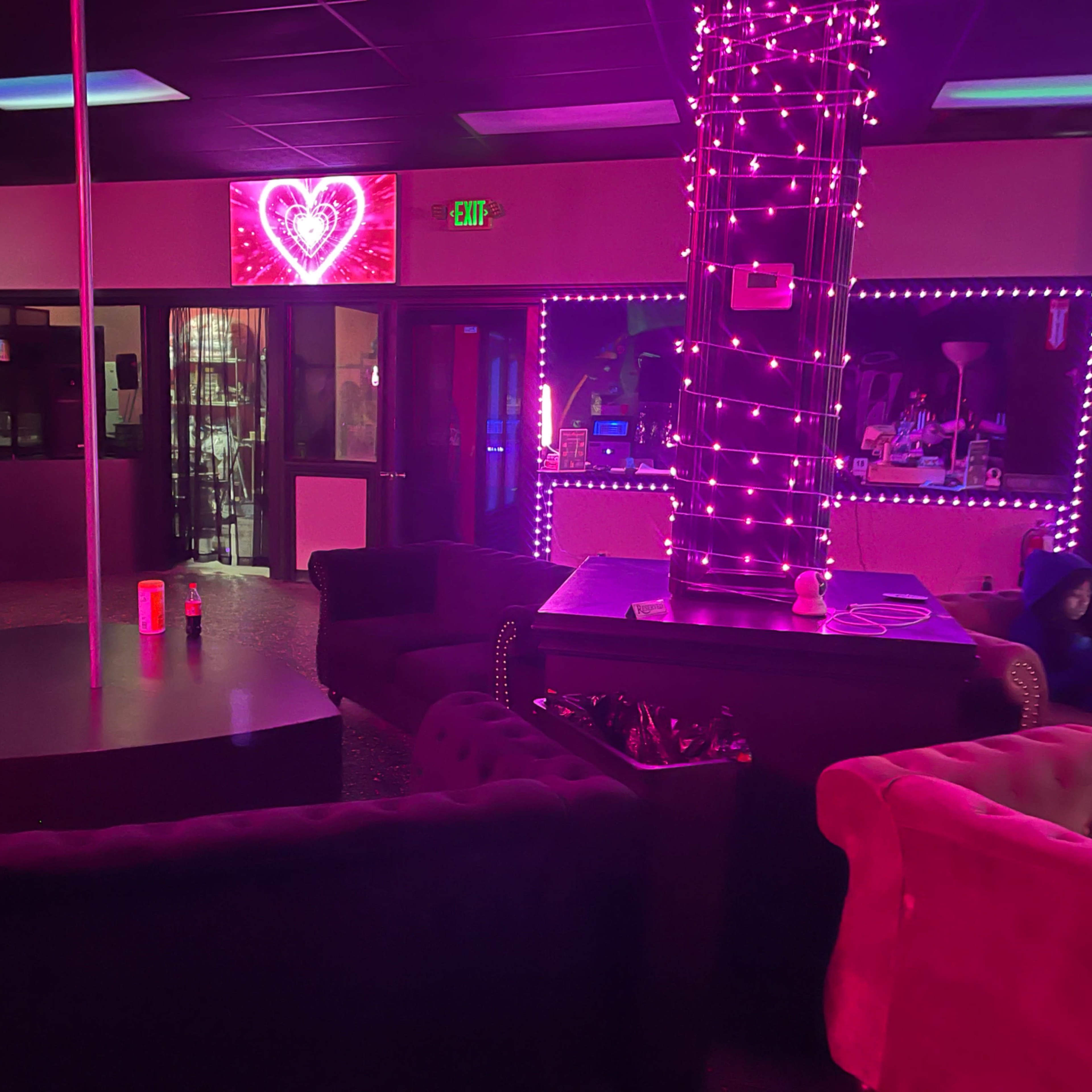 A dimly lit lounge with pink and purple lighting, featuring plush seating arranged around a central pole and illuminated decorative elements.
