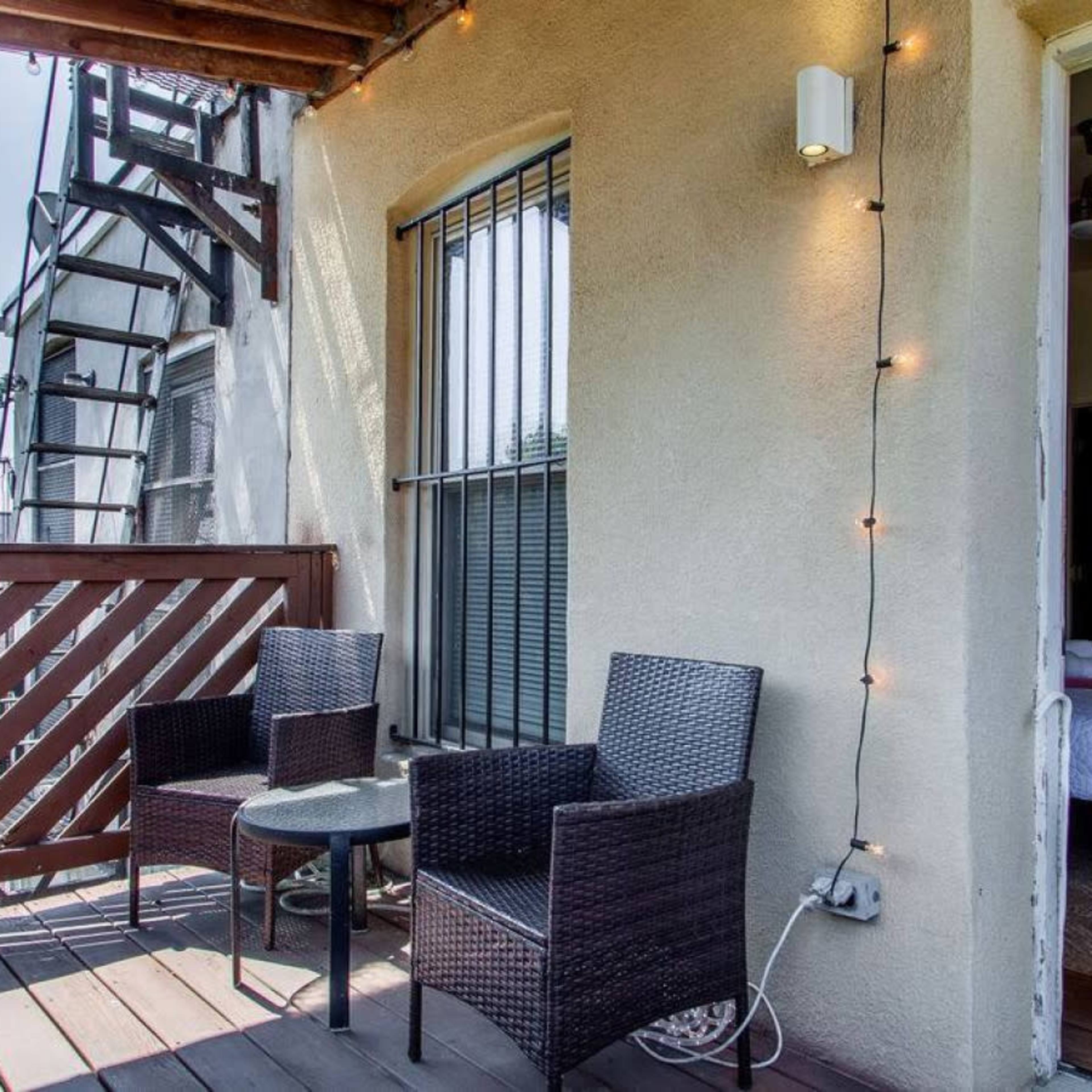 A small balcony features two wicker chairs and a table, with a doorway leading inside a room.