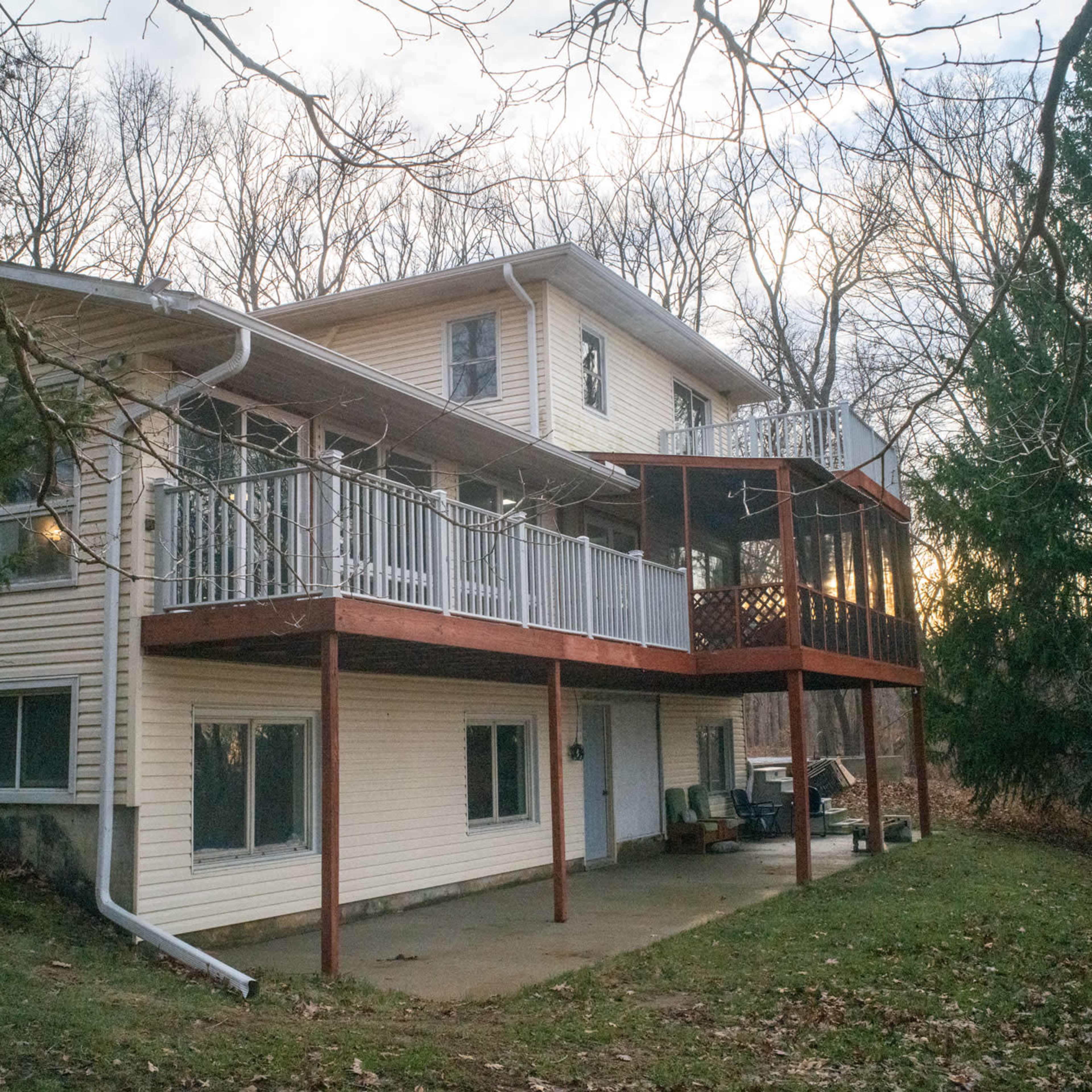 A two-story house features a large deck with a railing extending off the back, surrounded by trees and a grassy area.