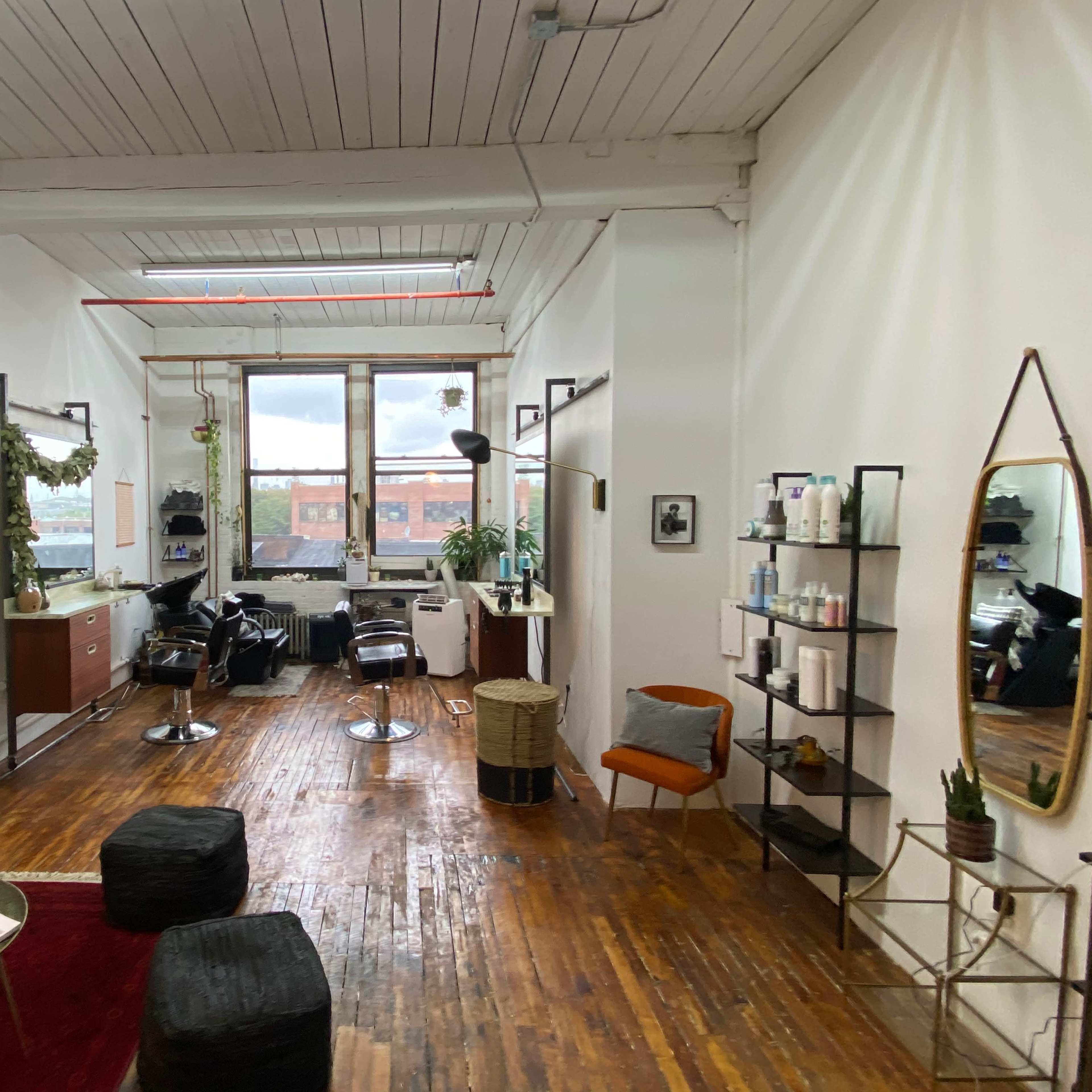 A well-lit hair salon with wooden floors, a large window, and several styling stations equipped with chairs and mirrors.