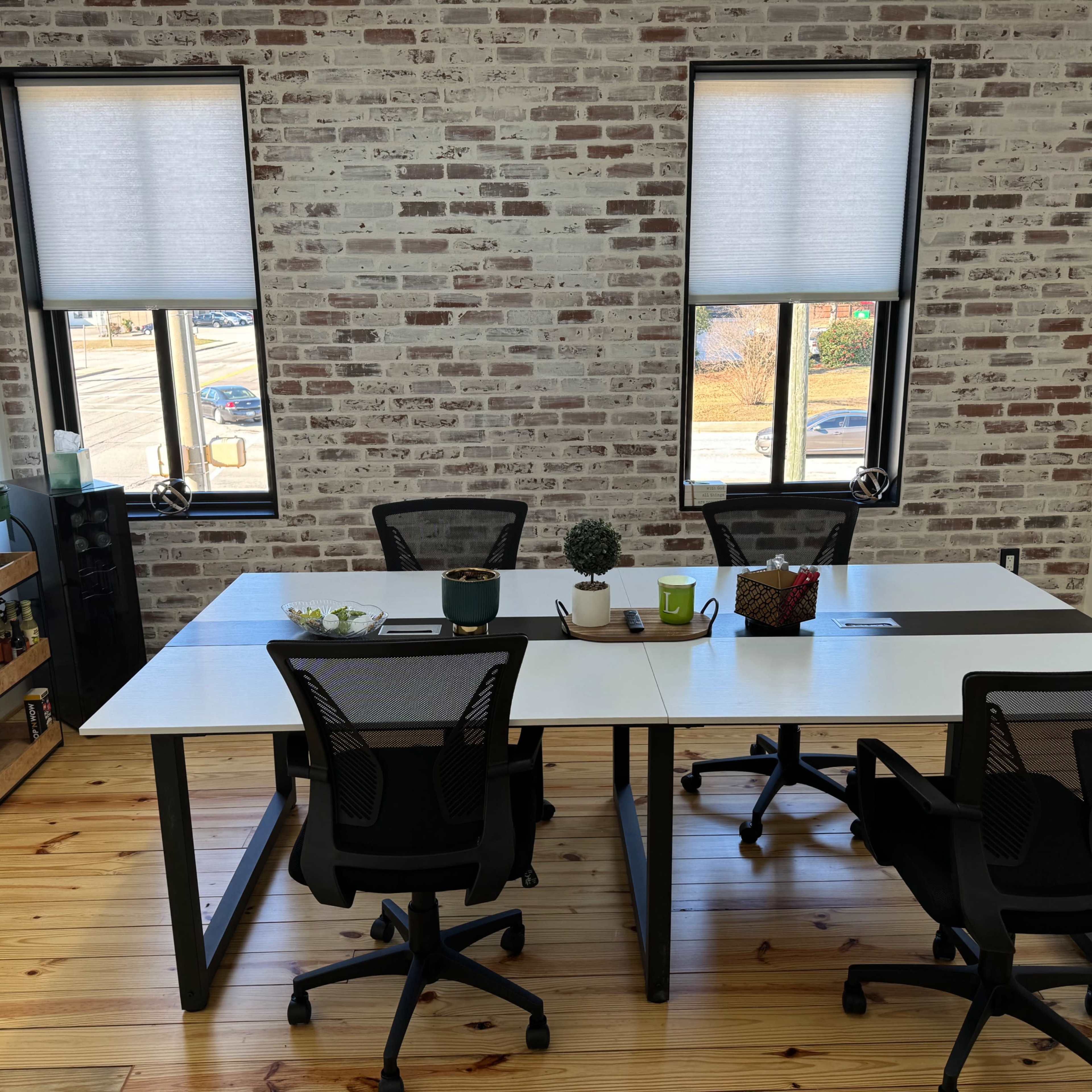 A modern conference room features a large table surrounded by black office chairs and two windows with blinds, set against a brick wall.
